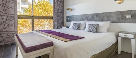 A modern hotel room with a large bed, a bedside table, a bench, a window with a view of buildings and autumn trees, and a wall-mounted light fixture.