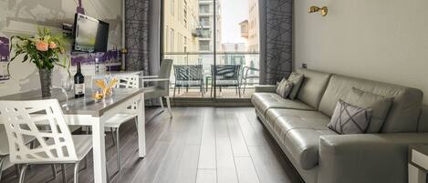 Un soggiorno moderno con un divano, un tavolo da pranzo con sedie, una TV e un balcone con vista sugli edifici.