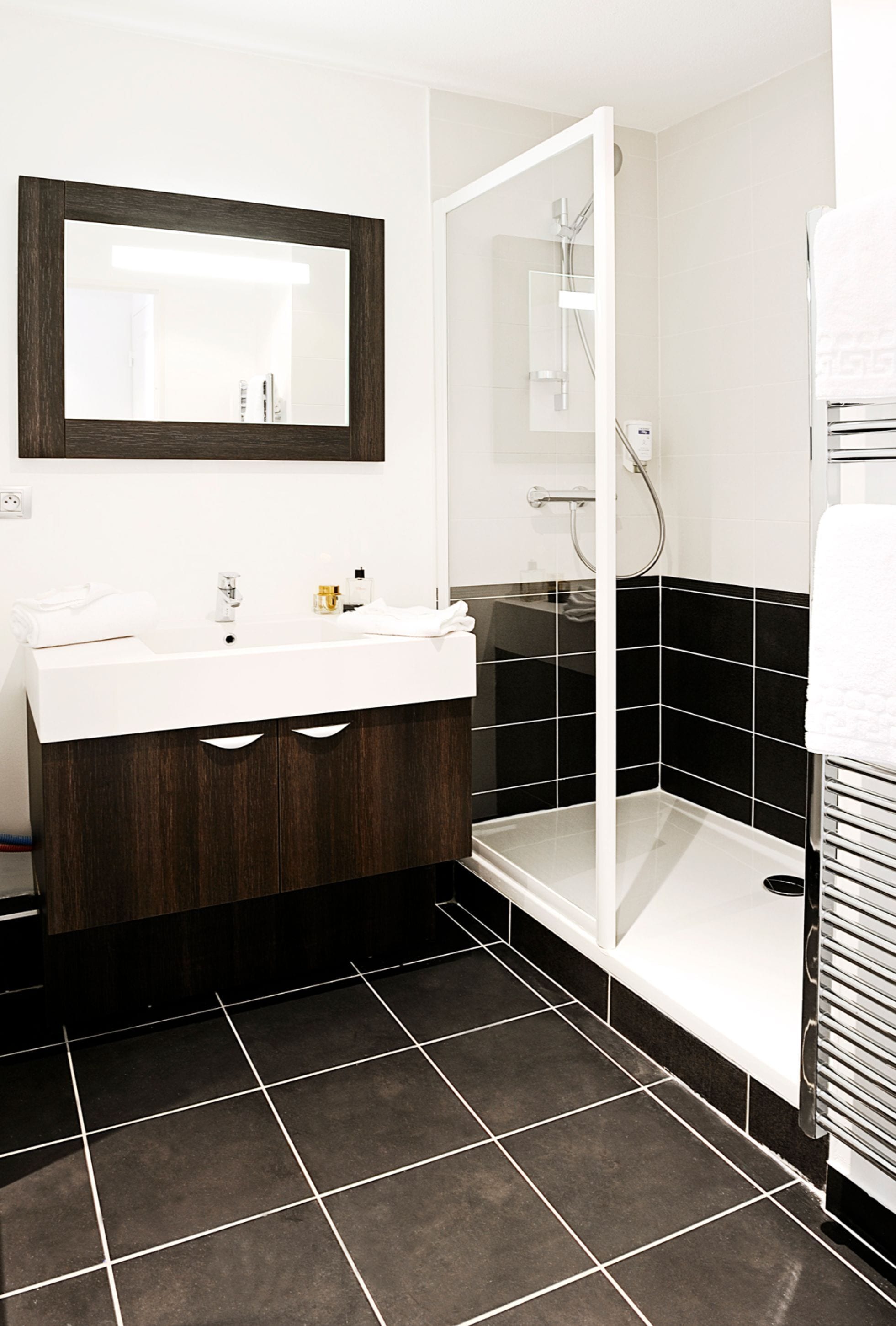 A modern bathroom with a white sink, dark wooden vanity, black and white tiled walls, a glass shower enclosure, and a white towel on the right.