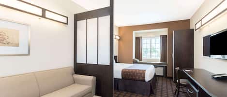 A hotel room with a sofa, a bed with a patterned bedspread, a desk with a chair, a television, and a window with curtains.