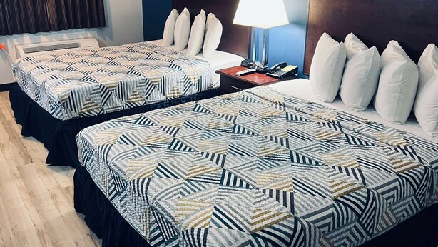 Two beds with patterned bedding, a bedside table with a lamp, and a wall-mounted air conditioner.