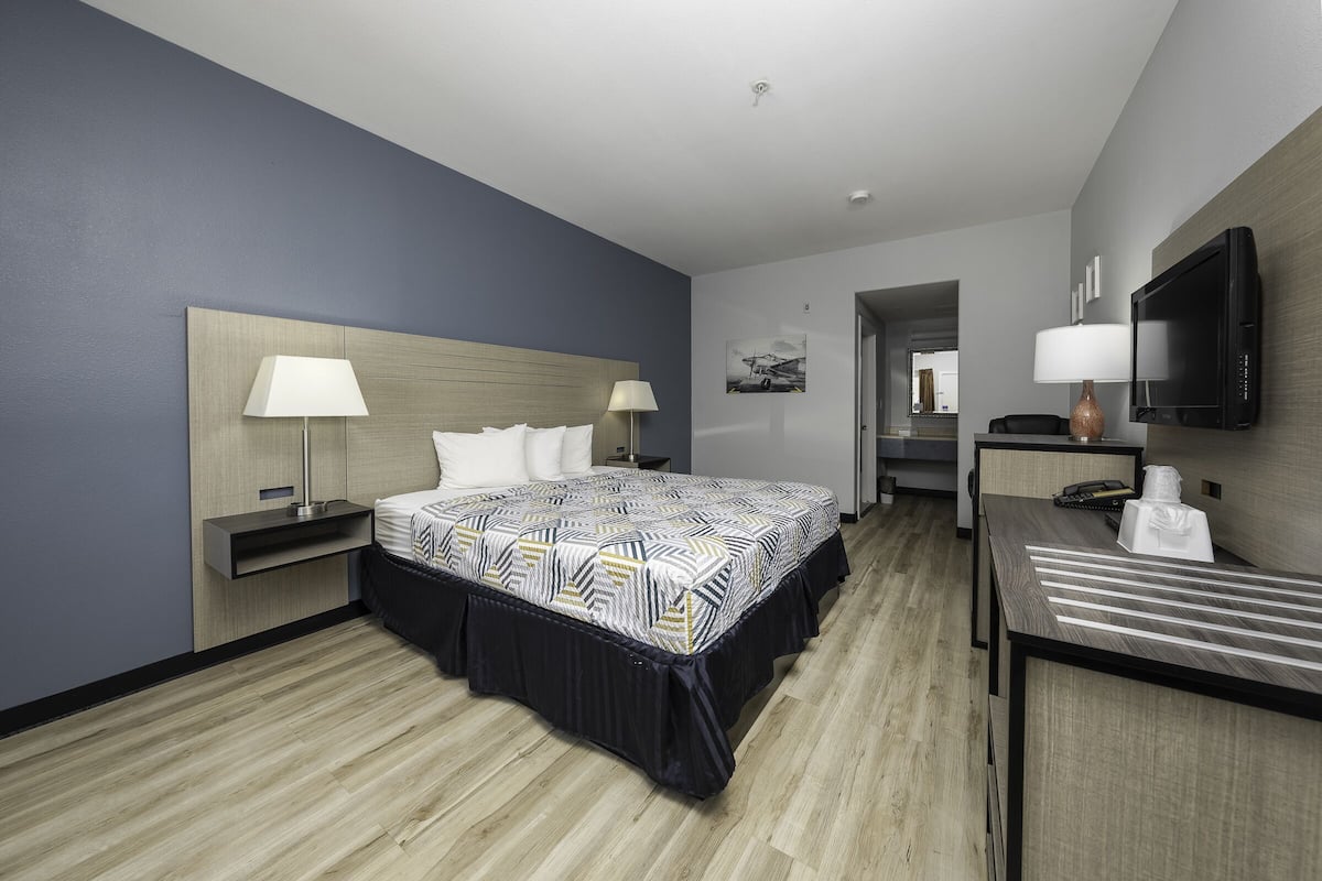 A modern hotel room with a large bed, two bedside tables with lamps, a desk with a chair, a flat-screen TV, and a wooden floor.