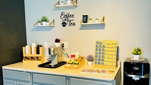 A kitchenette with a coffee station, a water dispenser, and a wall-mounted shelf with plants and a coffee-themed sign.