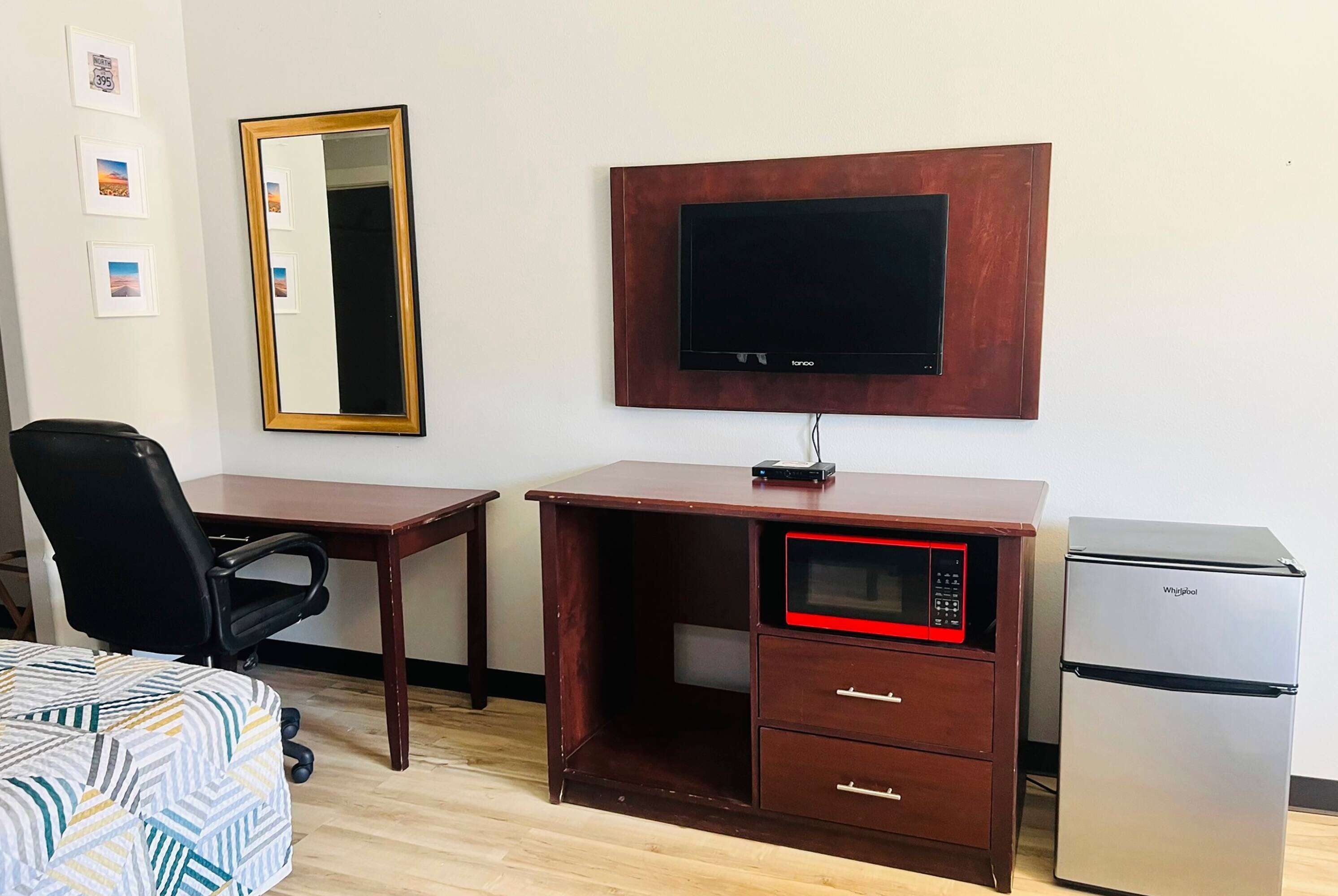 A compact hotel room with a wooden desk, a black office chair, a small refrigerator, a microwave, a flat-screen TV, and a mirror.