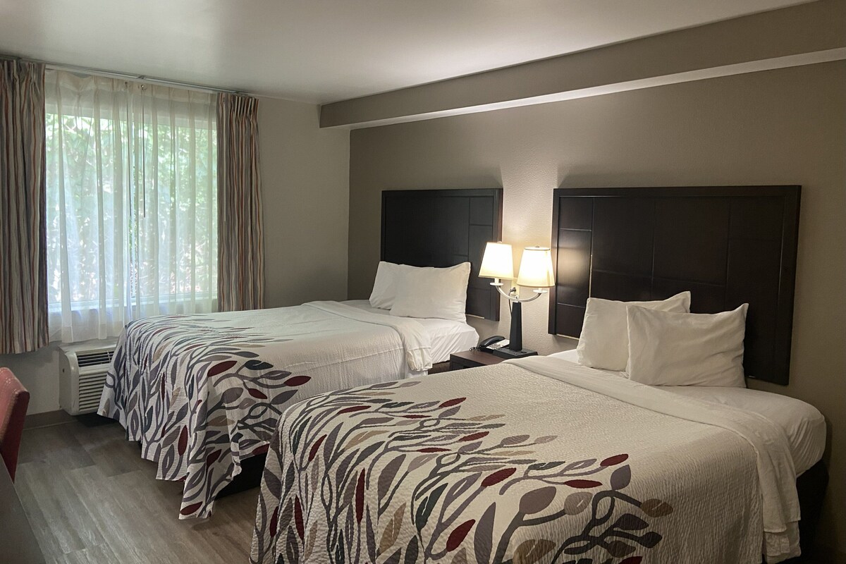 A hotel room with two beds, a nightstand with a lamp, and a window with curtains.