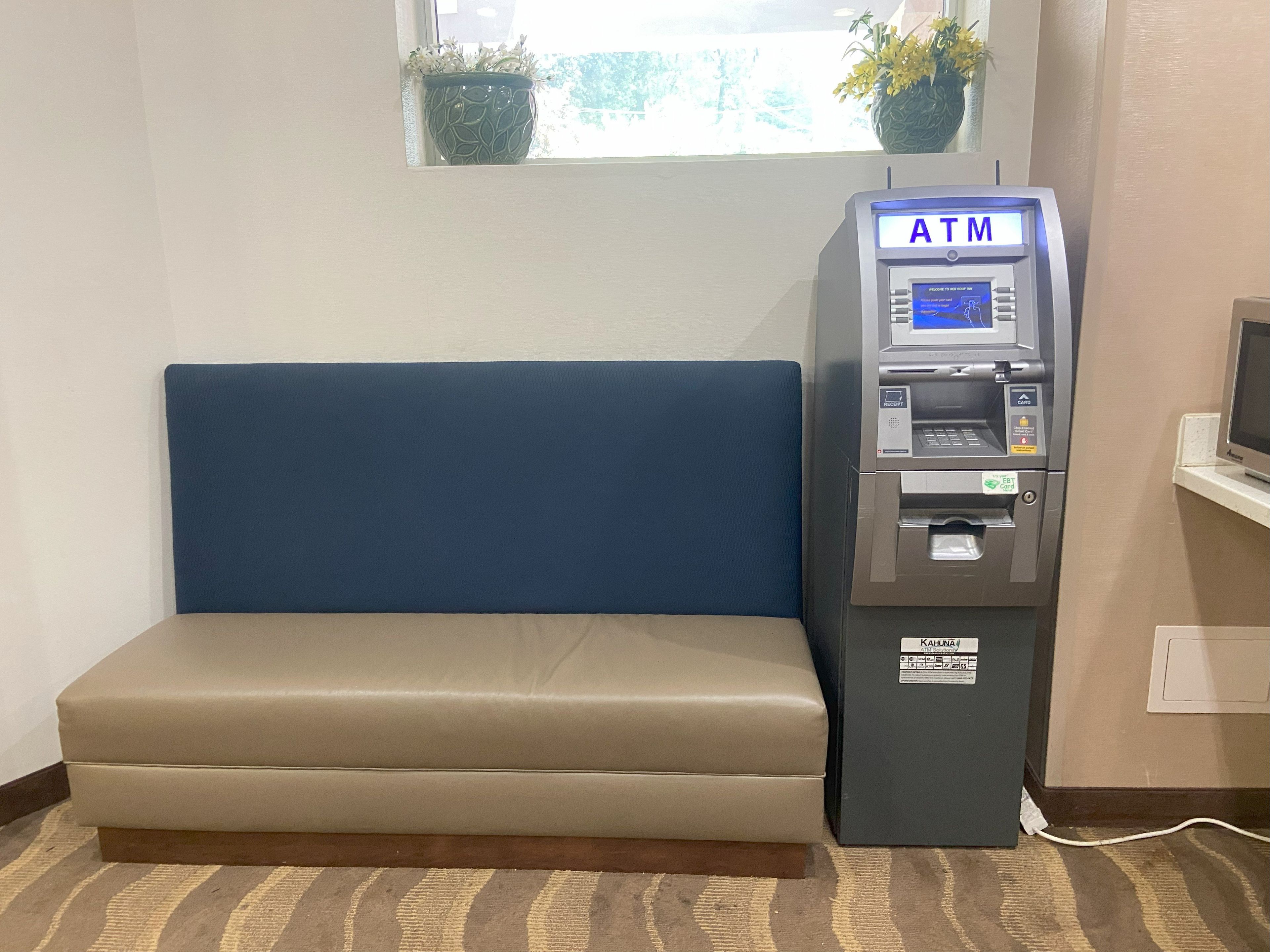 Un cajero automático al lado de un banco acolchado azul en una habitación con ventana y una planta.