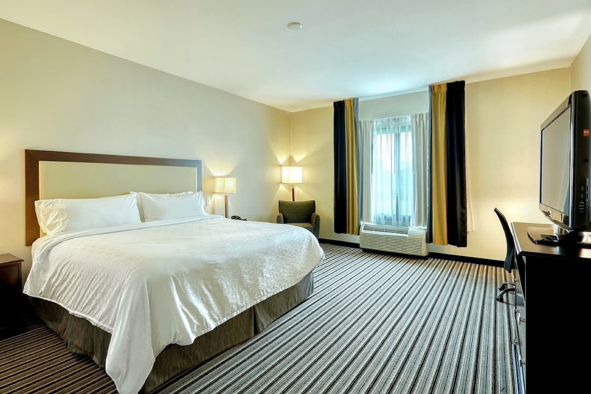Ein Hotelzimmer mit einem großen Bett, einem Schreibtisch mit Fernseher, einem Sessel und einem Fenster mit Vorhängen.