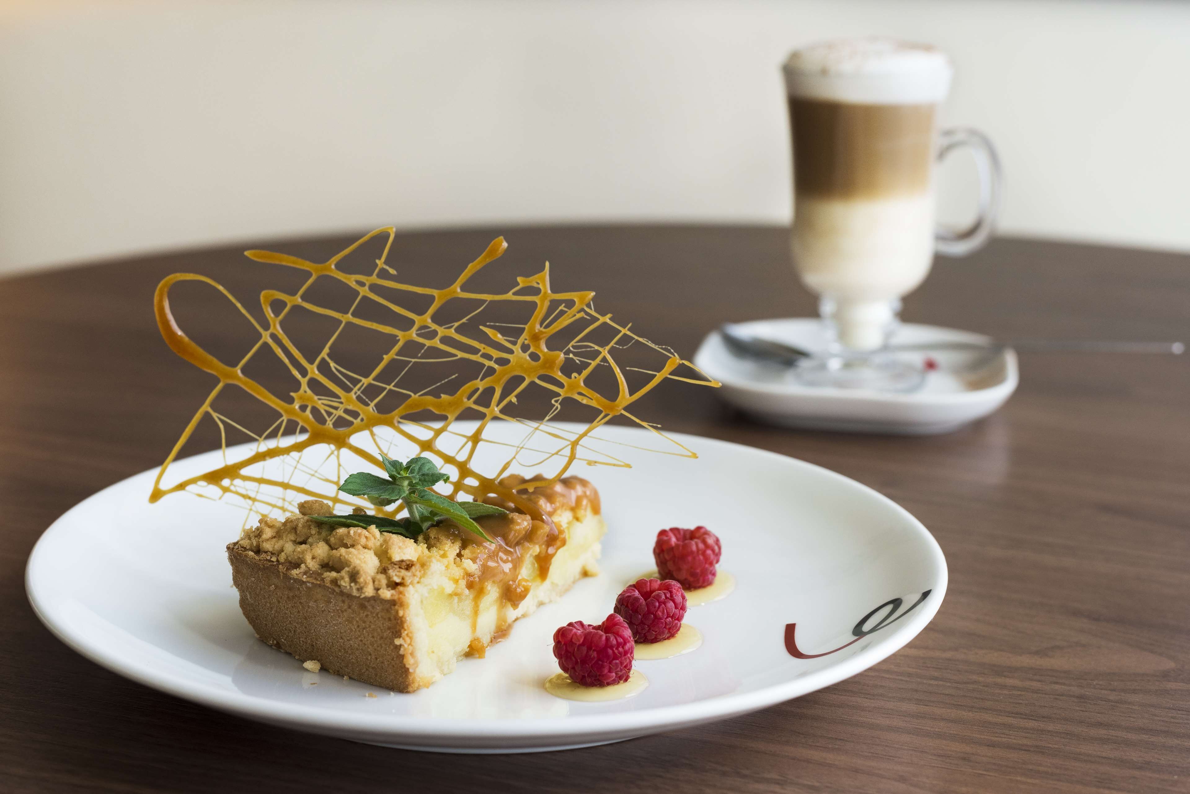 Un plato de postre con una porción de pastel, frambuesas y un hilo de caramelo sobre un plato blanco.