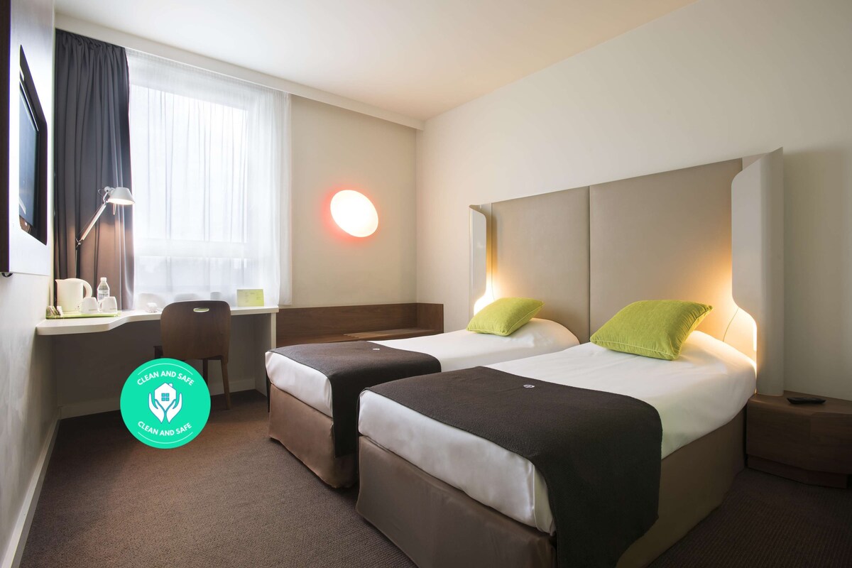 Habitación de hotel con dos camas, un escritorio, una silla y una ventana con cortinas.