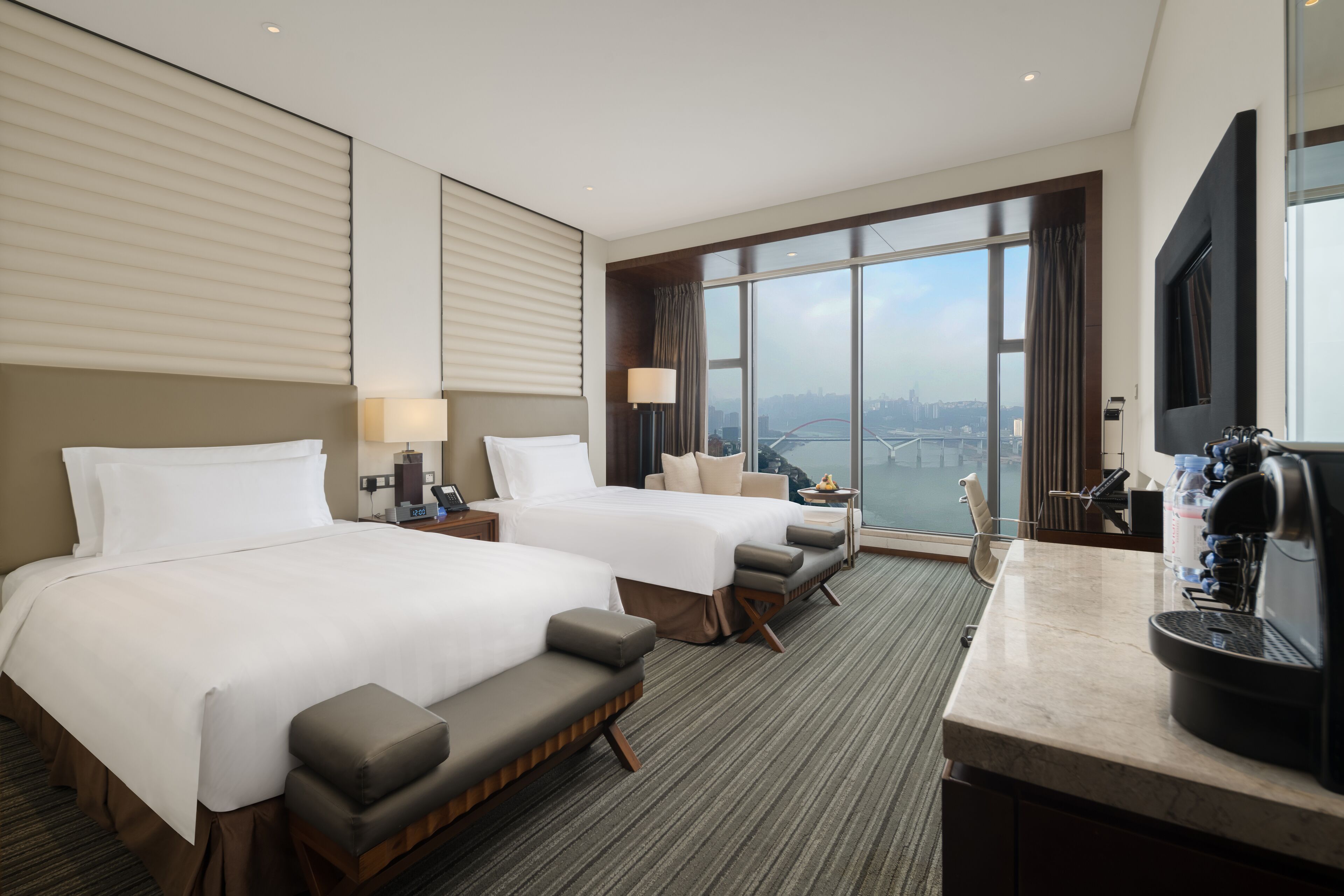 A hotel room with two beds, a sofa, a desk with a coffee machine, and a large window with a city view.