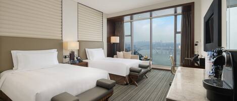 A hotel room with two beds, a sofa, a desk with a coffee machine, and a large window with a city view.