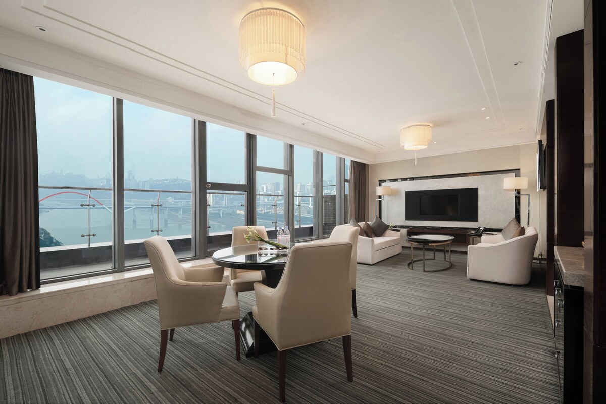 A modern hotel room with a large window offering a city view, a flat-screen TV, and a seating area with a round table.