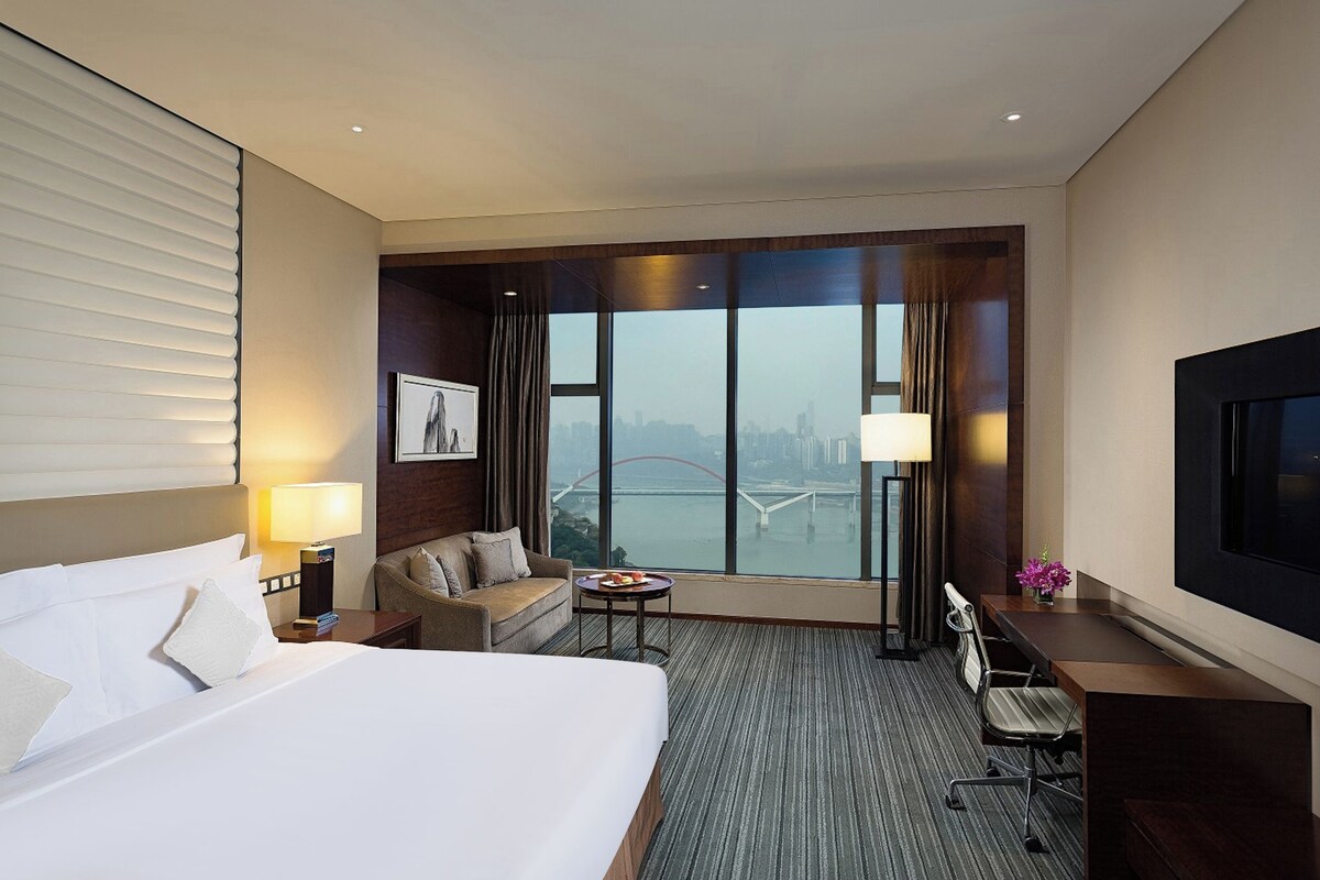 A hotel room with a large bed, a desk, a chair, a sofa, a TV, and a view of a cityscape through large windows.