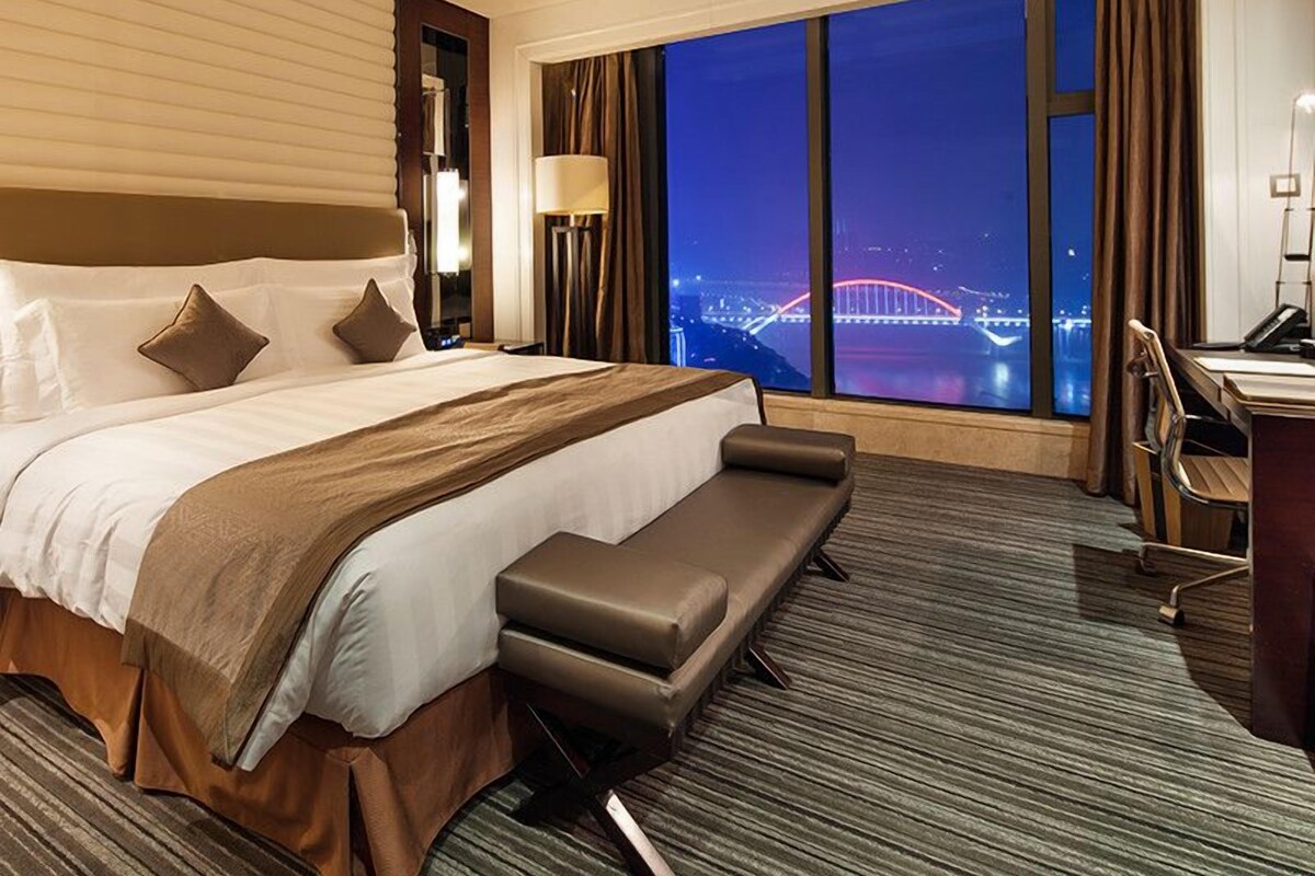 A hotel room with a large bed, a desk, and a view of a bridge at night.