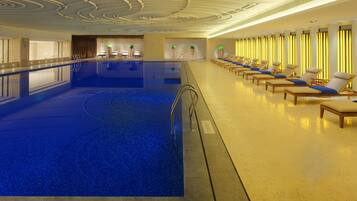Una amplia piscina cubierta con superficie de agua cristalina, rodeada de sillones de descanso y una zona de recepción con mostrador y sillas.