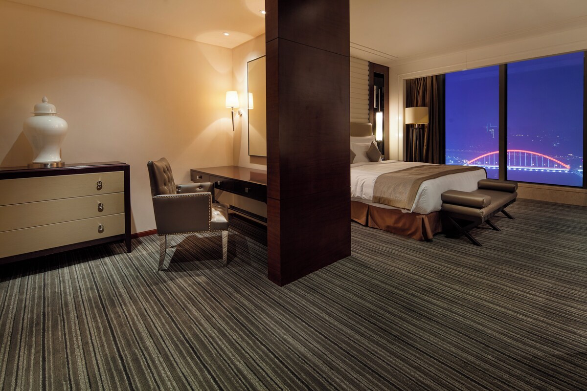 Habitación de hotel con cama, escritorio, silla, cómoda, lámpara y un amplio ventanal con vista nocturna.
