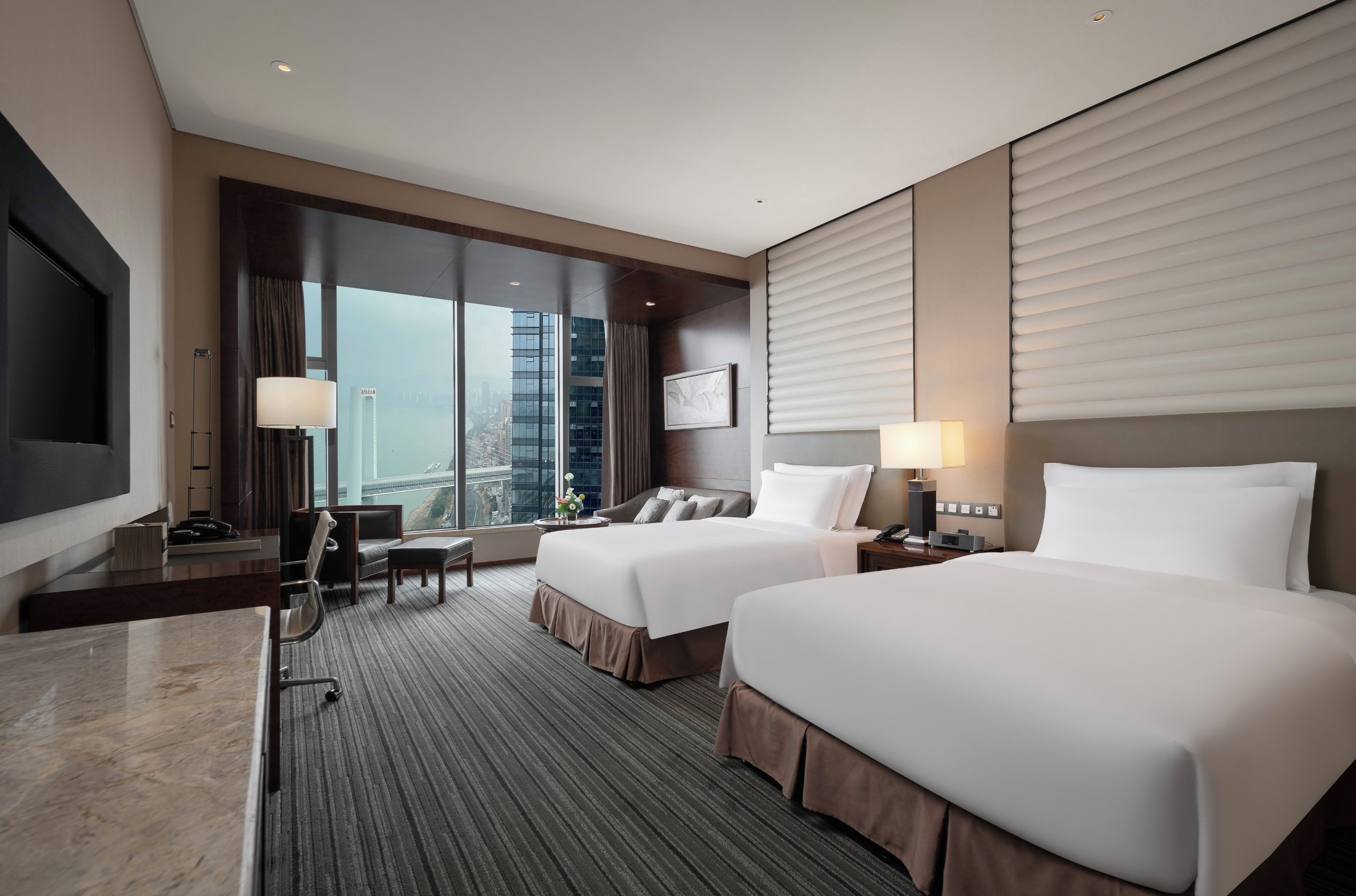 A hotel room with two beds, a desk, a chair, a TV, and a large window with a city view.