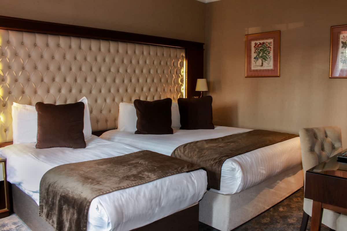 A hotel room with two beds, a tufted headboard, a nightstand with a lamp, and framed floral artwork on the walls.