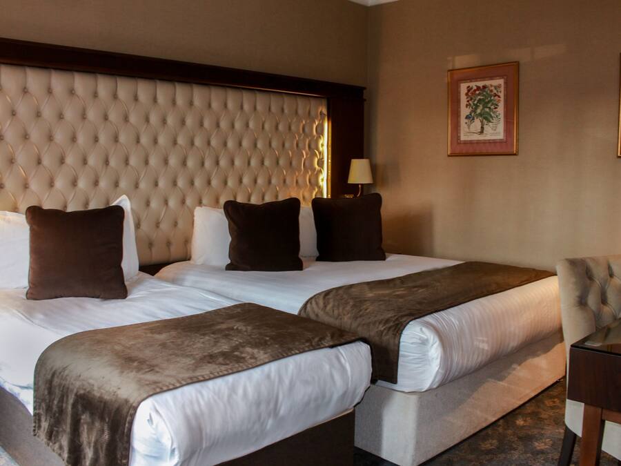 A hotel room with two beds, a tufted headboard, a nightstand with a lamp, and framed floral artwork on the walls.