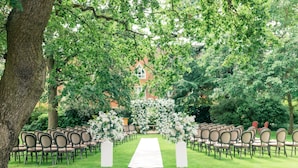 An outdoor wedding venue with a white aisle, chairs arranged on both sides, and a backdrop of trees and a building. - Warren House (Kingston Upon Thames)