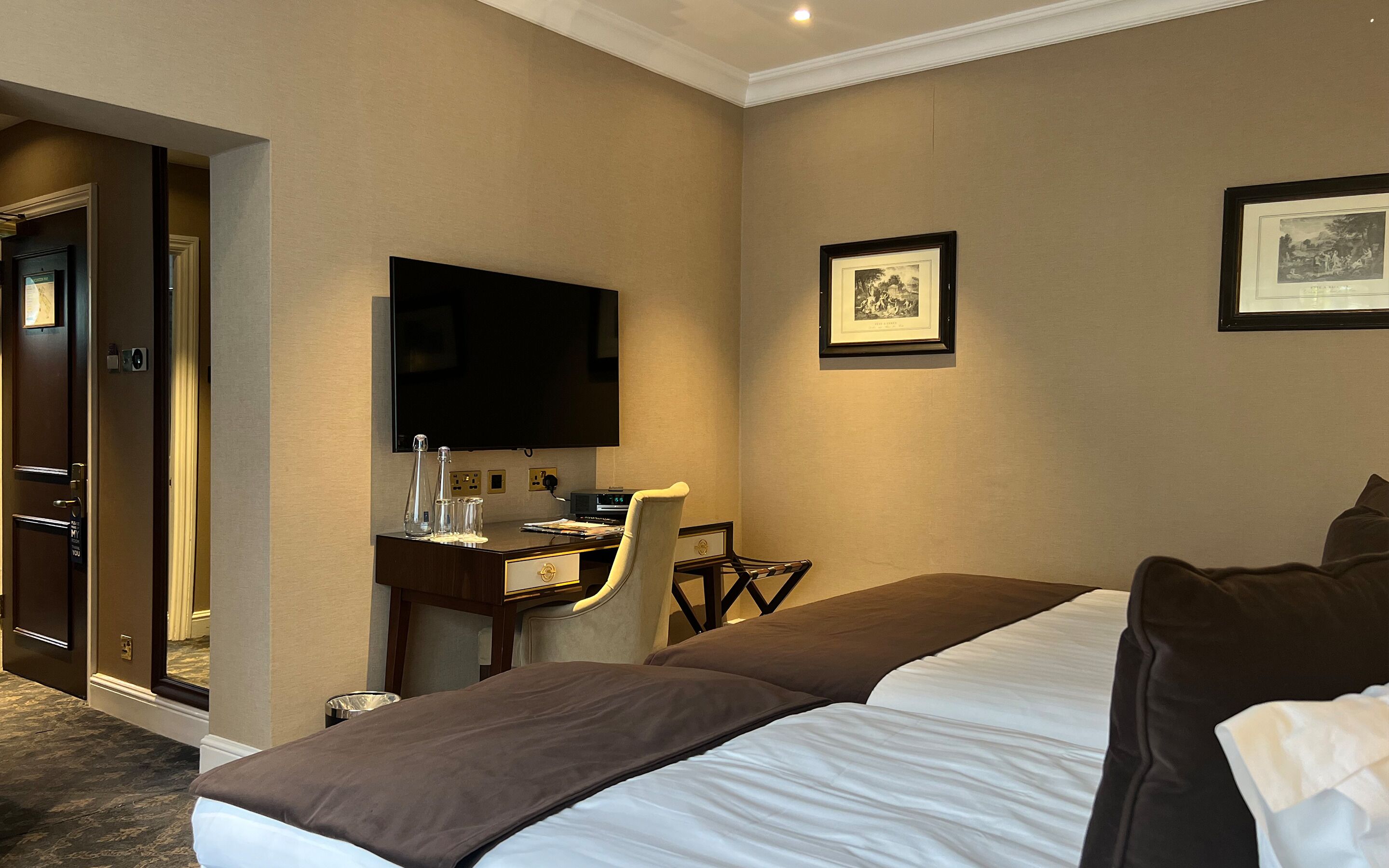 Ein Hotelzimmer mit Bett, Schreibtisch mit Stuhl, Flachbildfernseher und zwei gerahmten Bildern an der Wand.