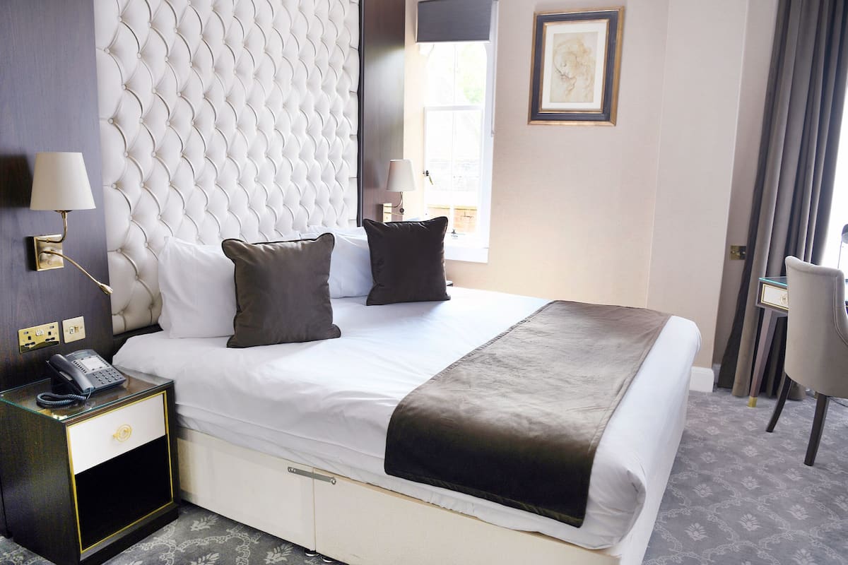 A hotel room with a tufted headboard, a bedside telephone, a desk with a chair, and a window with curtains.