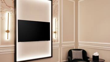A modern hotel room with a large flat-screen TV mounted on the wall, a comfortable black chair, a small round table with a tissue box, and a bed with a beige blanket.