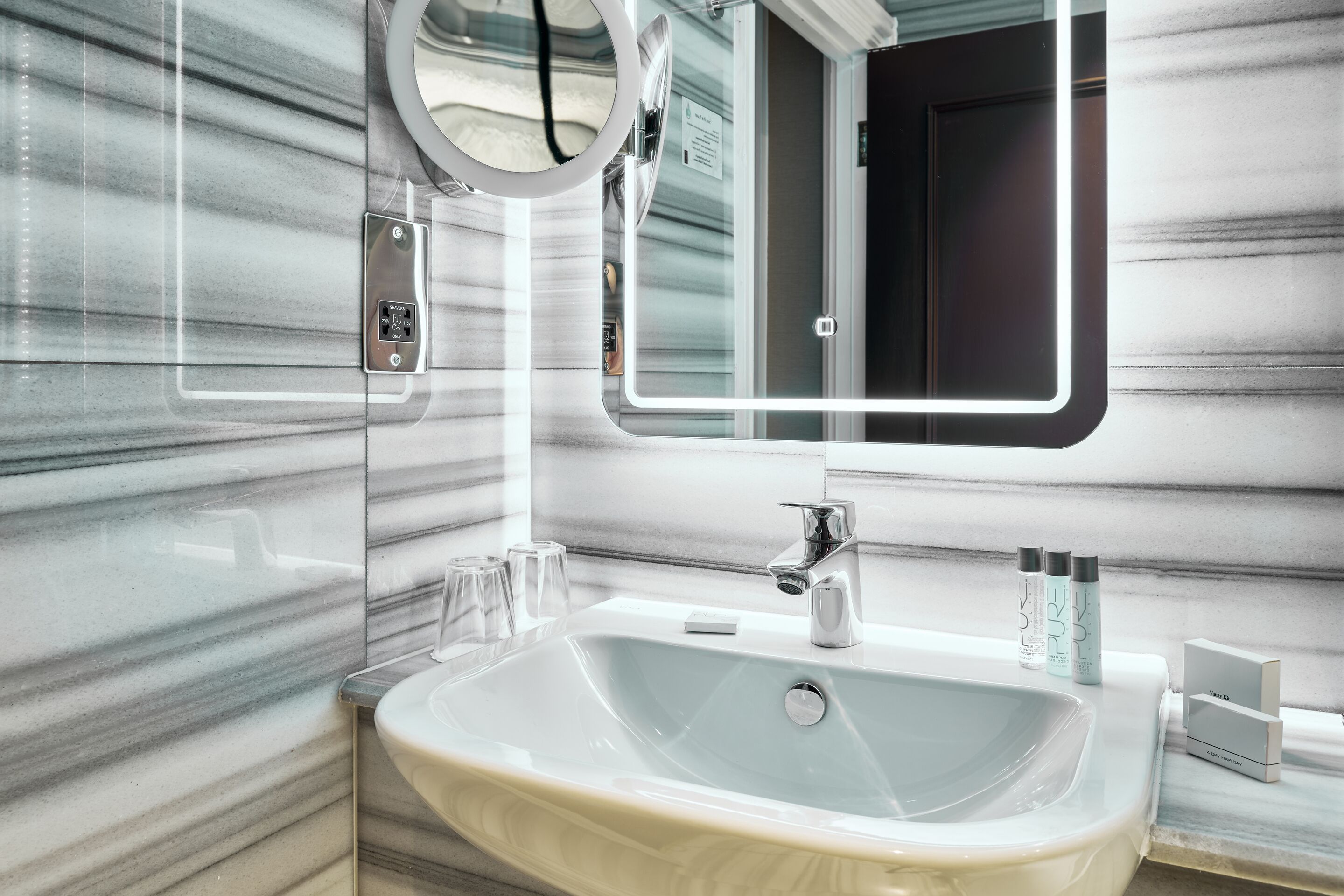 A modern bathroom with a marble backsplash, a large sink, a mirror with a light, and toiletries on the counter.