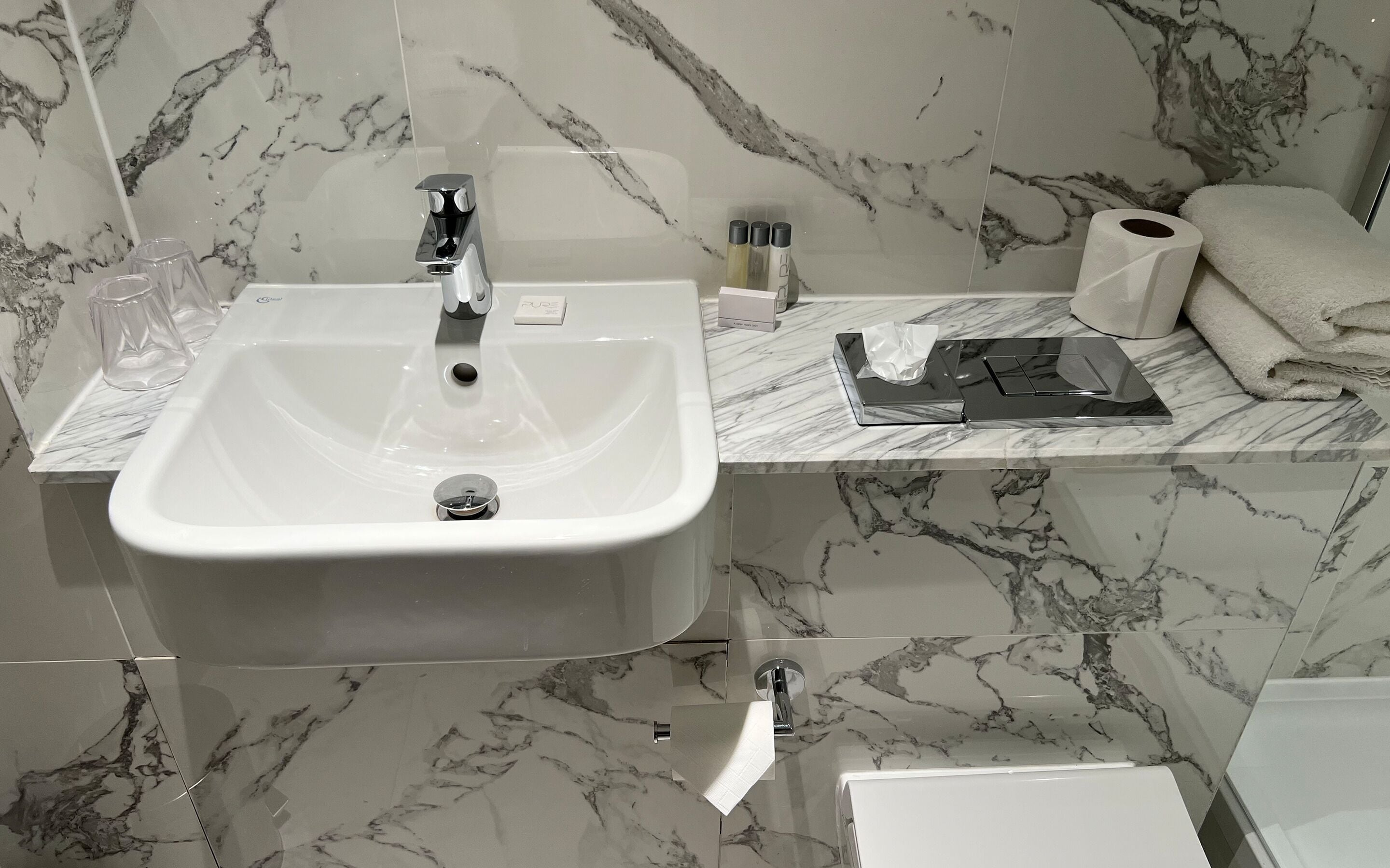 Un baño con un lavamanos blanco, un espejo, un inodoro y una pared con azulejos de mármol.