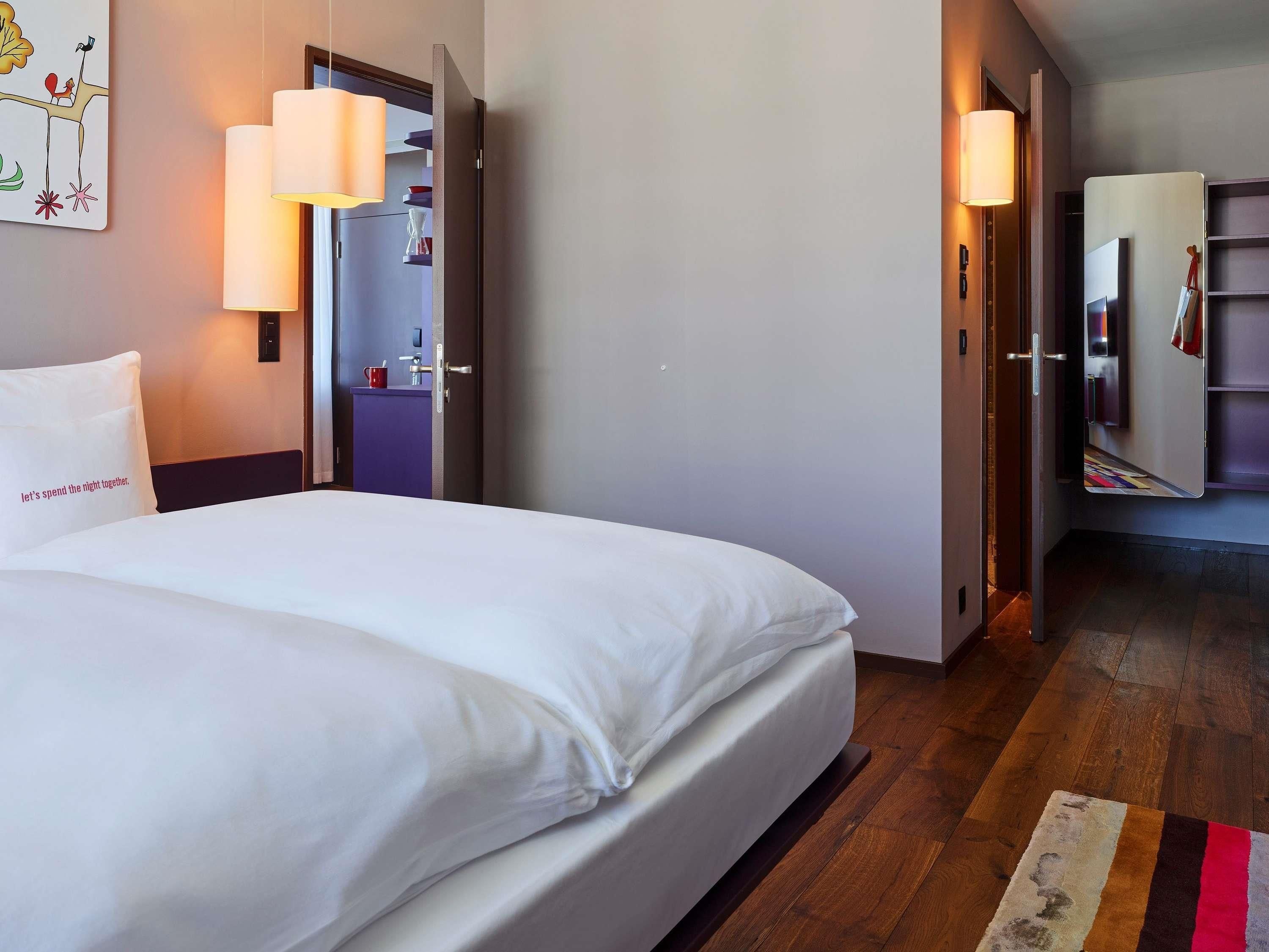 A hotel room with a bed, bedside lamps, a nightstand, a wall-mounted artwork, and a wooden floor.