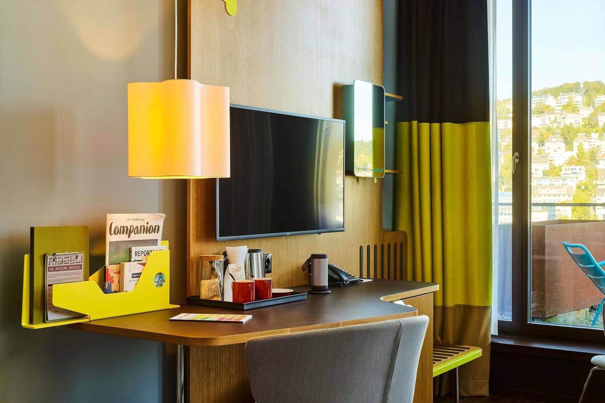 A hotel room with a desk, a flat-screen TV, a lamp, a chair, a window with curtains, and a view of buildings outside.