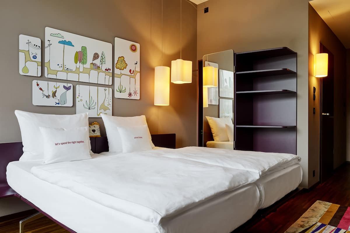 A modern hotel room with a large bed, white bedding, a bedside table with a lamp, a mirror, and artwork on the wall.