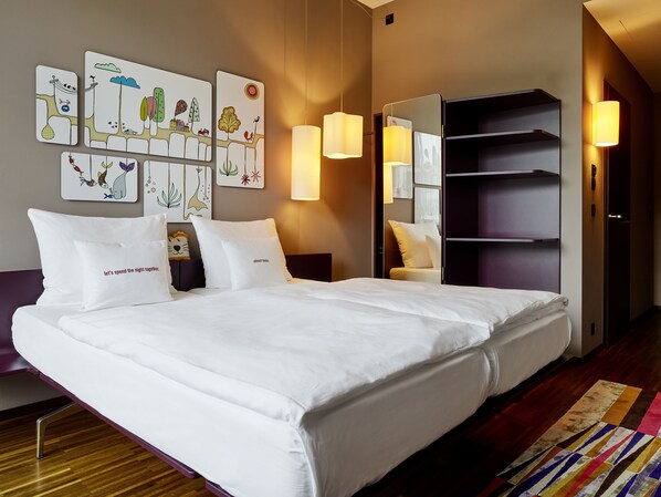 Una habitación de hotel moderna con una cama grande, ropa de cama blanca, una mesita de noche con lámpara, un espejo y una obra de arte en la pared.