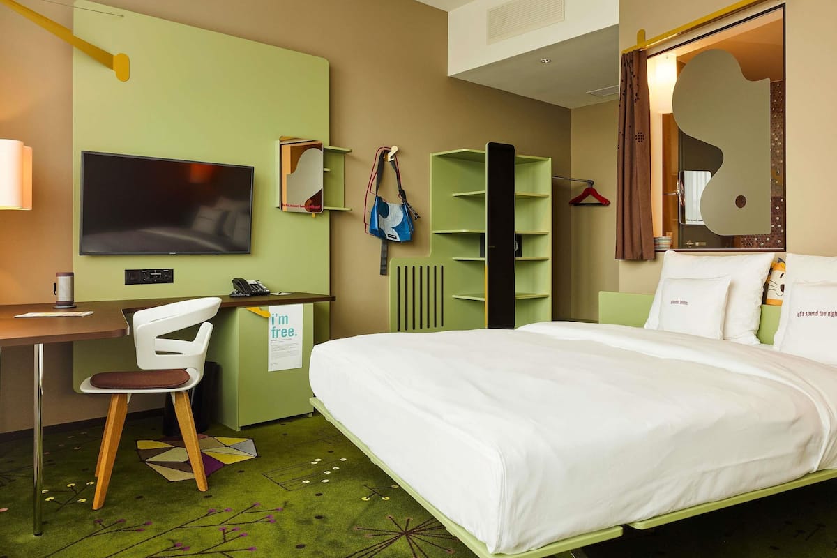 A hotel room with a bed, a desk with a chair, a TV, a wardrobe, and a mirror.