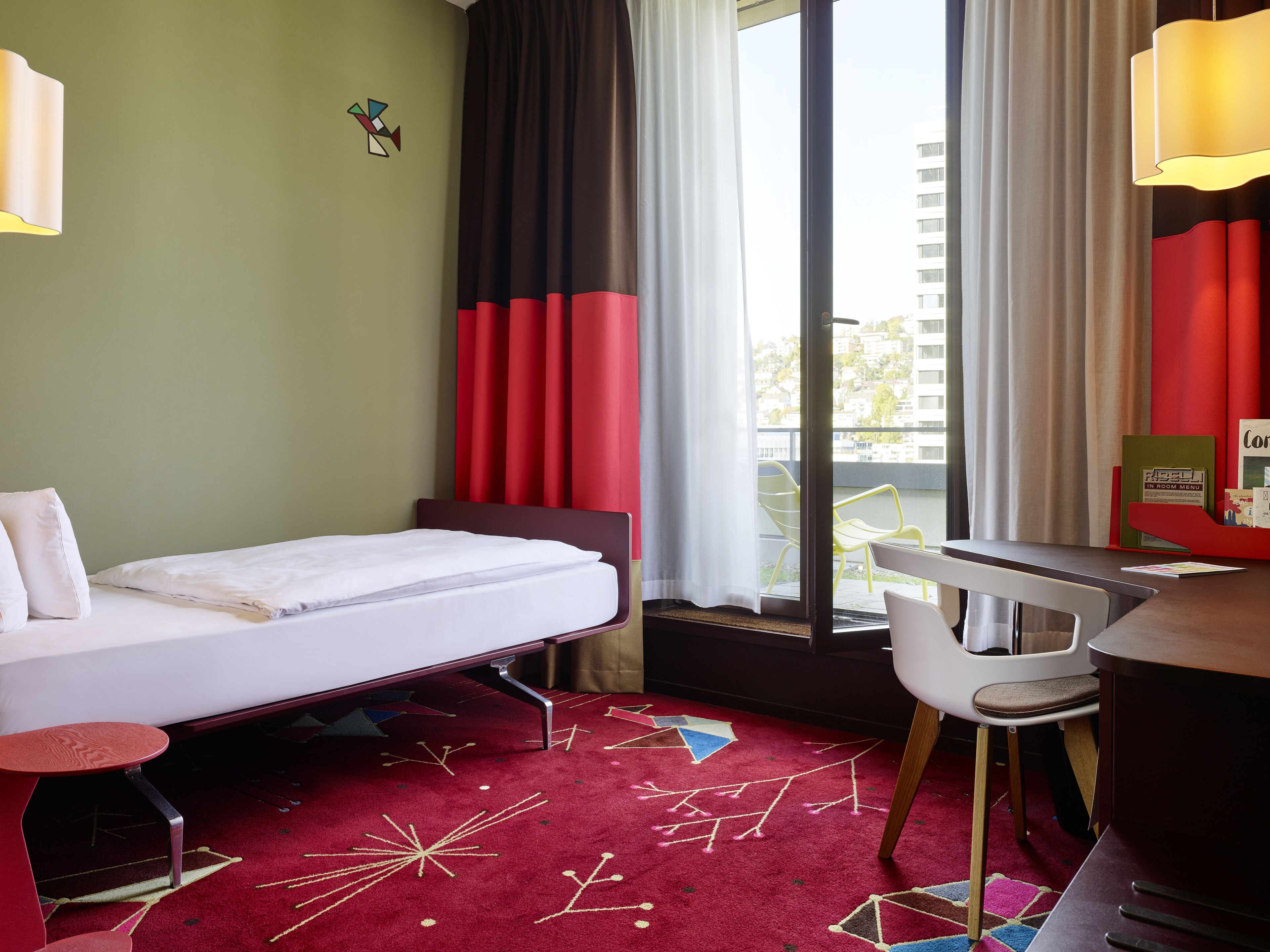 A hotel room with a bed, a desk, a chair, a red carpet with geometric patterns, and a view of buildings through the window.