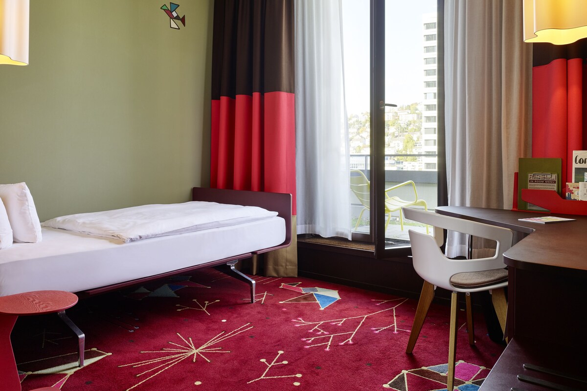 Une chambre d’hôtel avec un lit, un bureau, une chaise, une moquette rouge à motifs géométriques et une vue sur les bâtiments par la fenêtre.