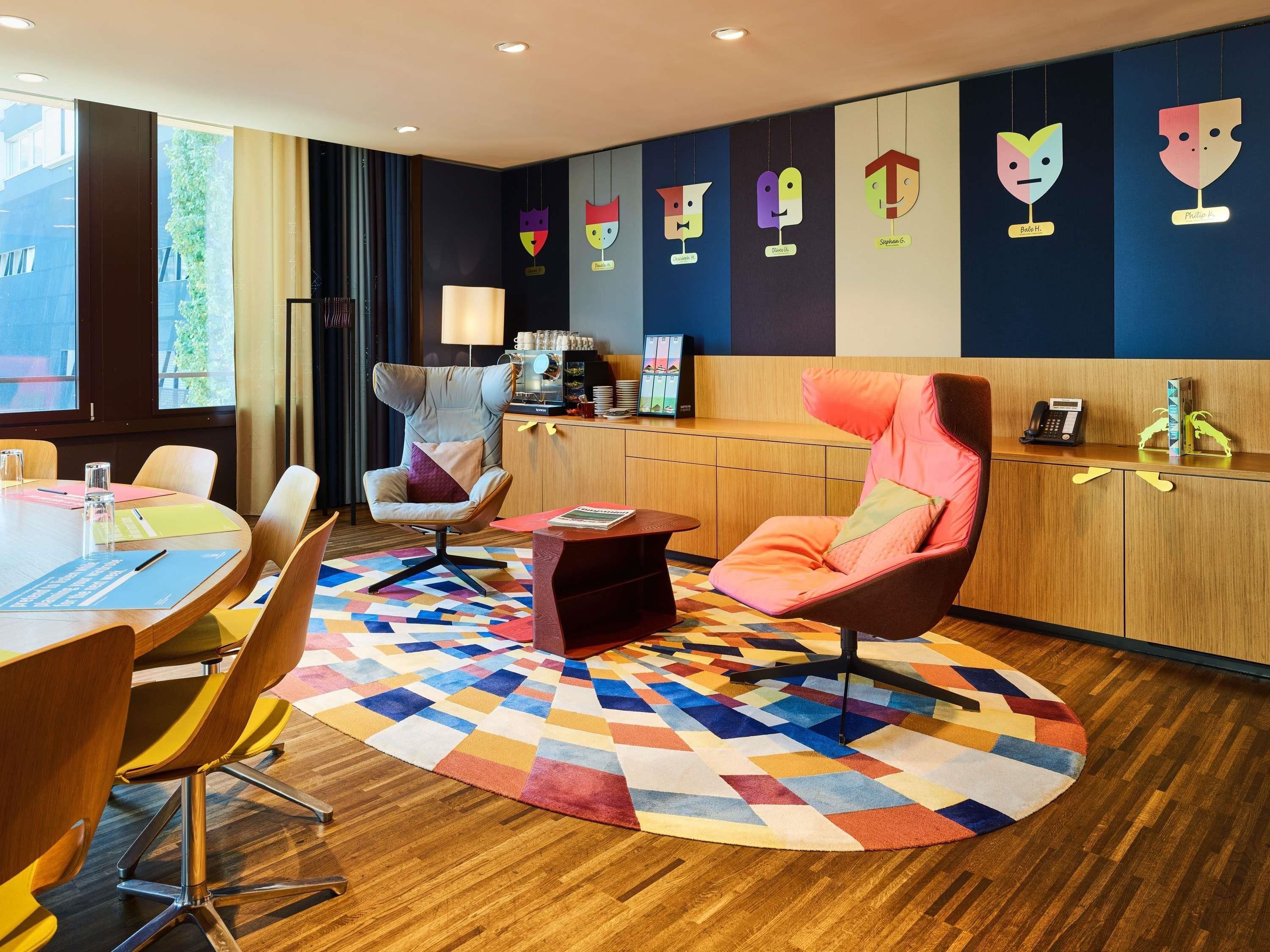 Una camera d'albergo moderna con un tavolo rotondo, sedute confortevoli, un tappeto decorato e una parete con opere d'arte colorate.