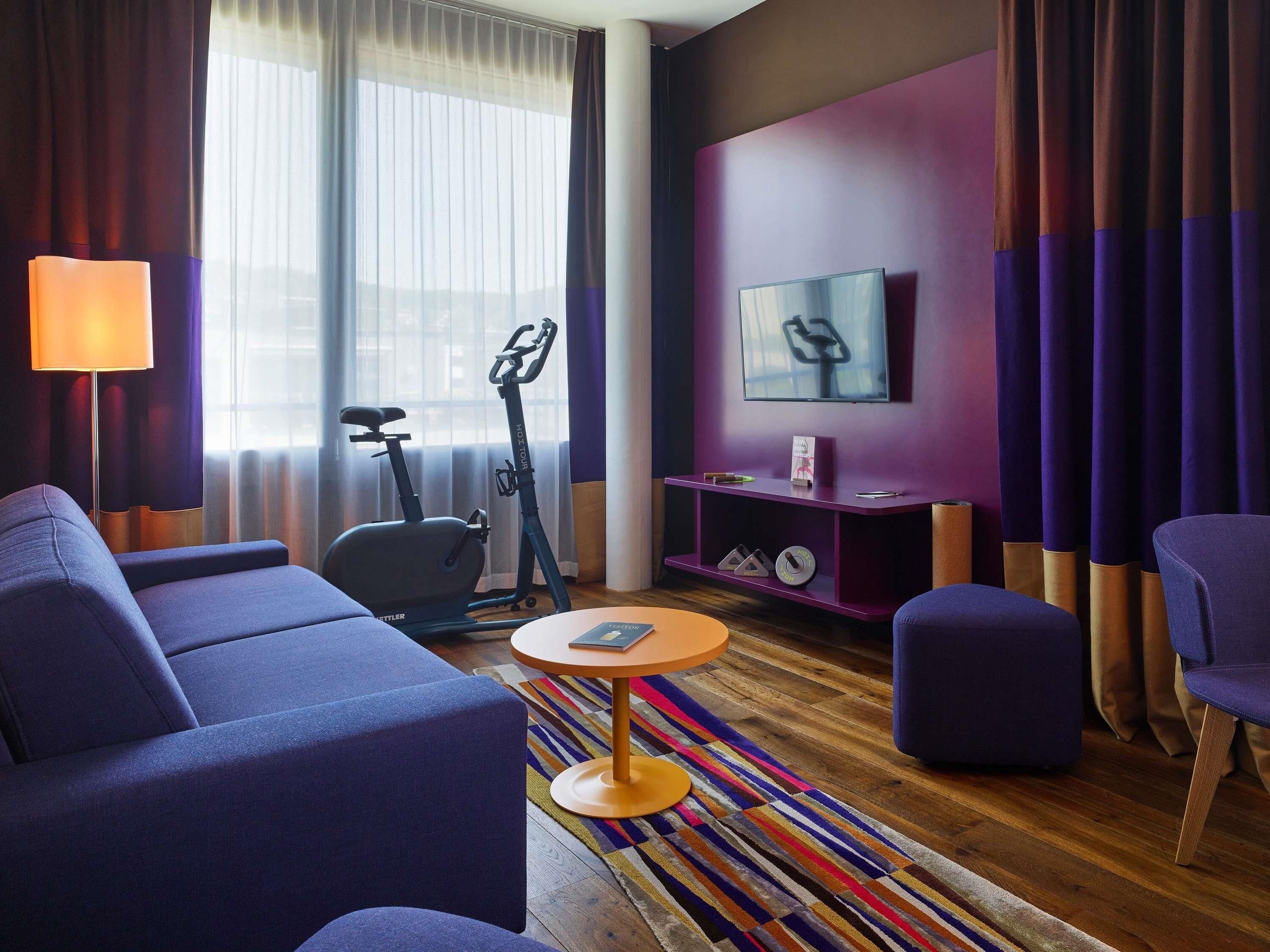 Habitación de hotel con sofá, una pequeña mesa redonda, televisor, cinta de correr y alfombra de colores.