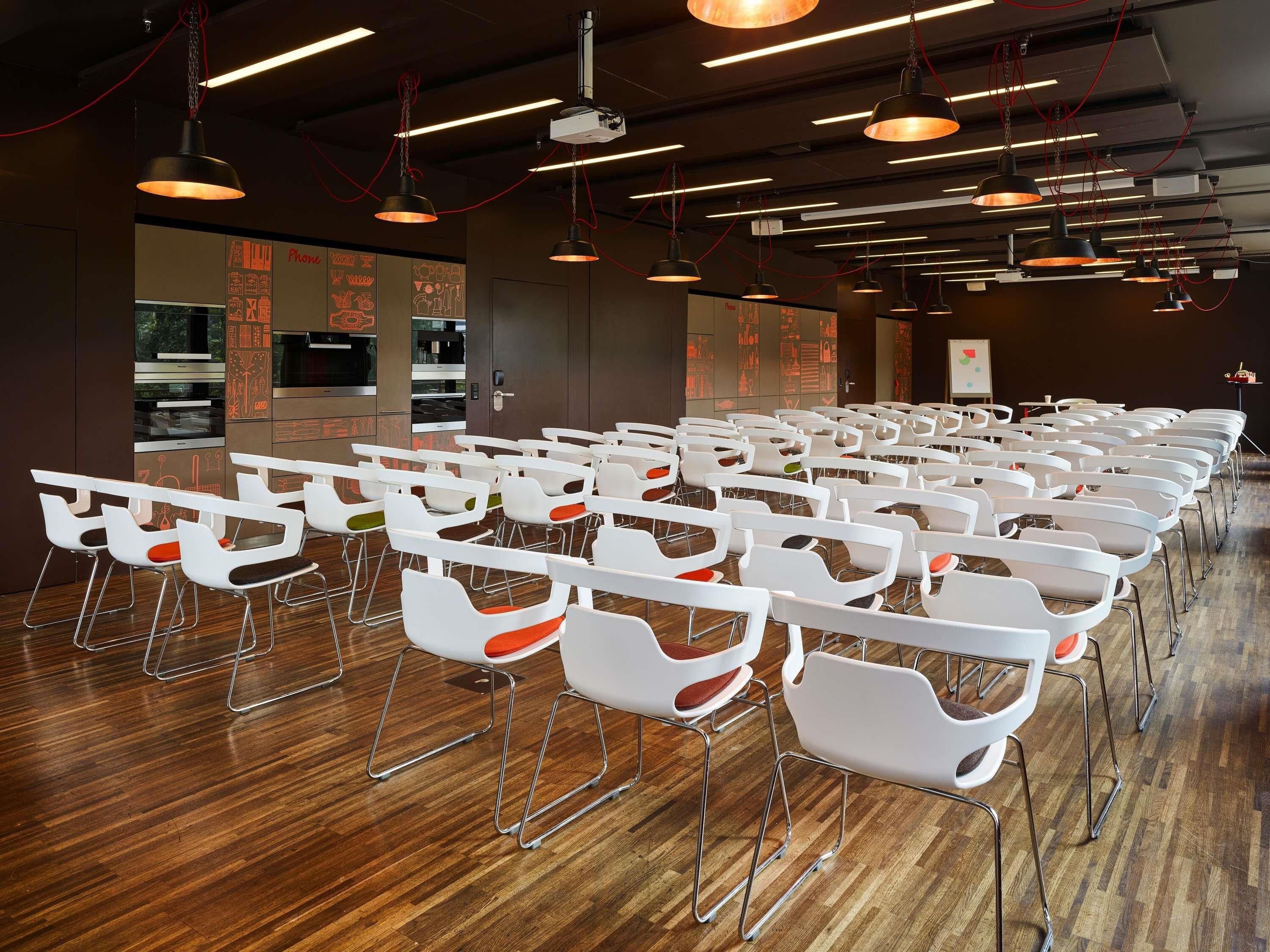 Une salle de conférence avec des rangées de chaises et de tables blanches, un plancher en bois et un éclairage moderne suspendu au plafond.