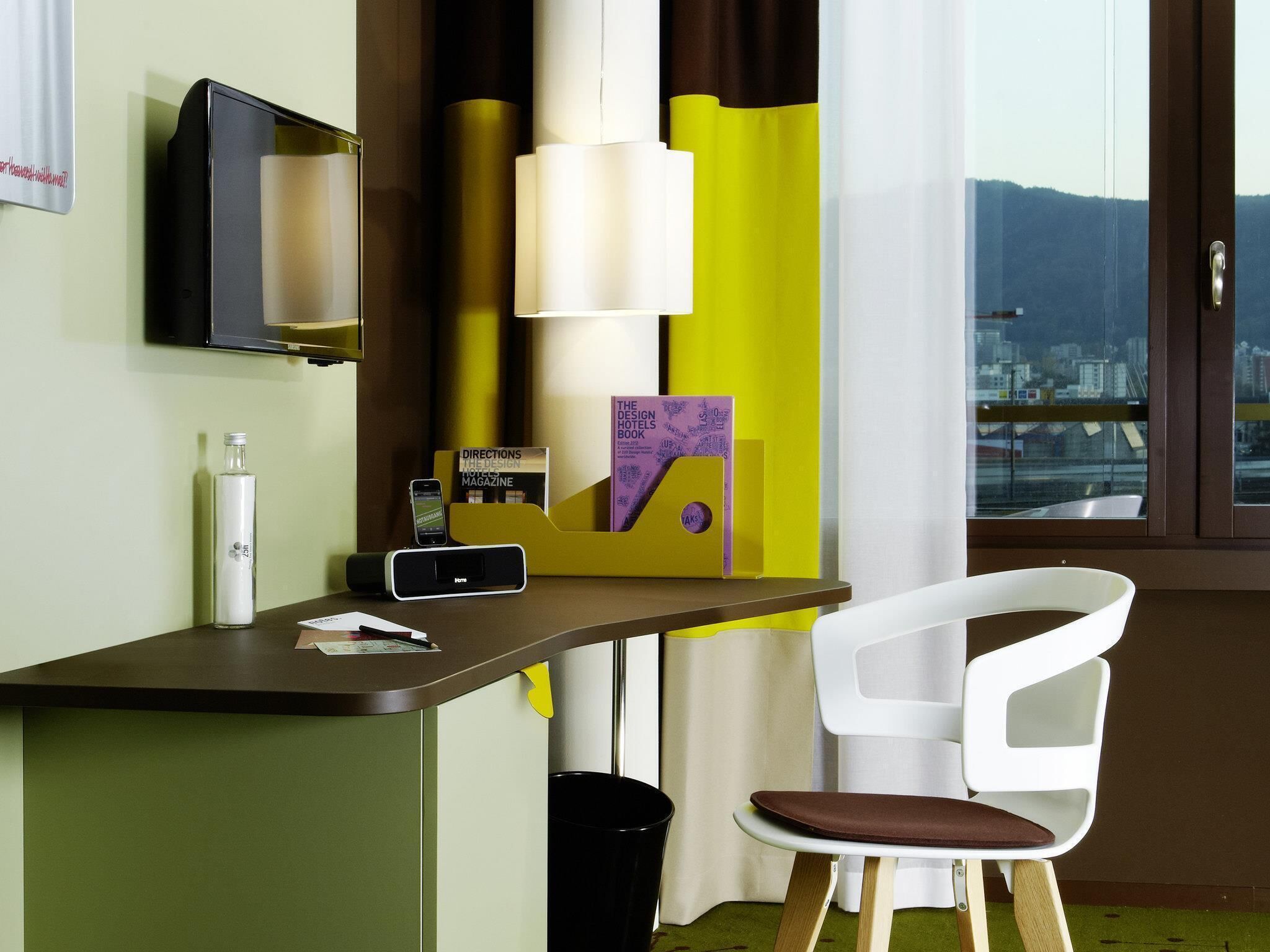 Hotelkamer met een bureau, een stoel, een televisie, een lamp, een boek, een mobiele telefoon en een raam met uitzicht op de stad.