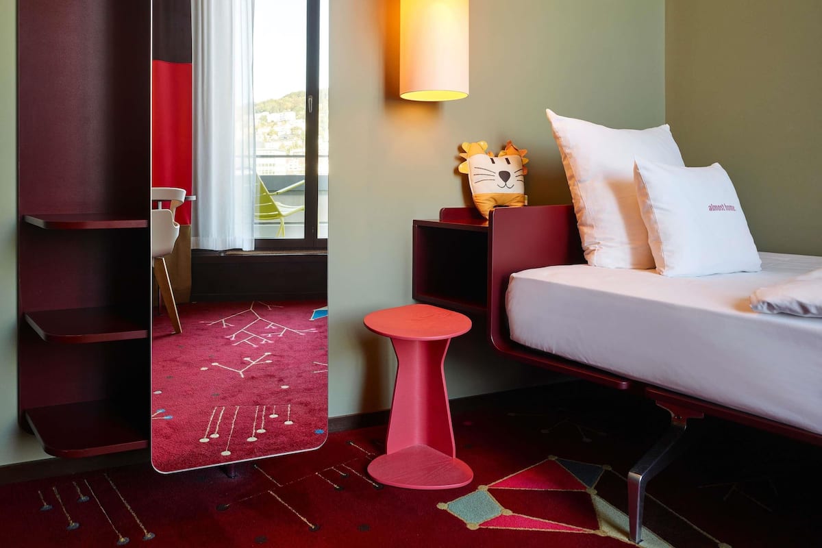 A hotel room with a bed, a red chair, a small table, a lamp, and a window with curtains.