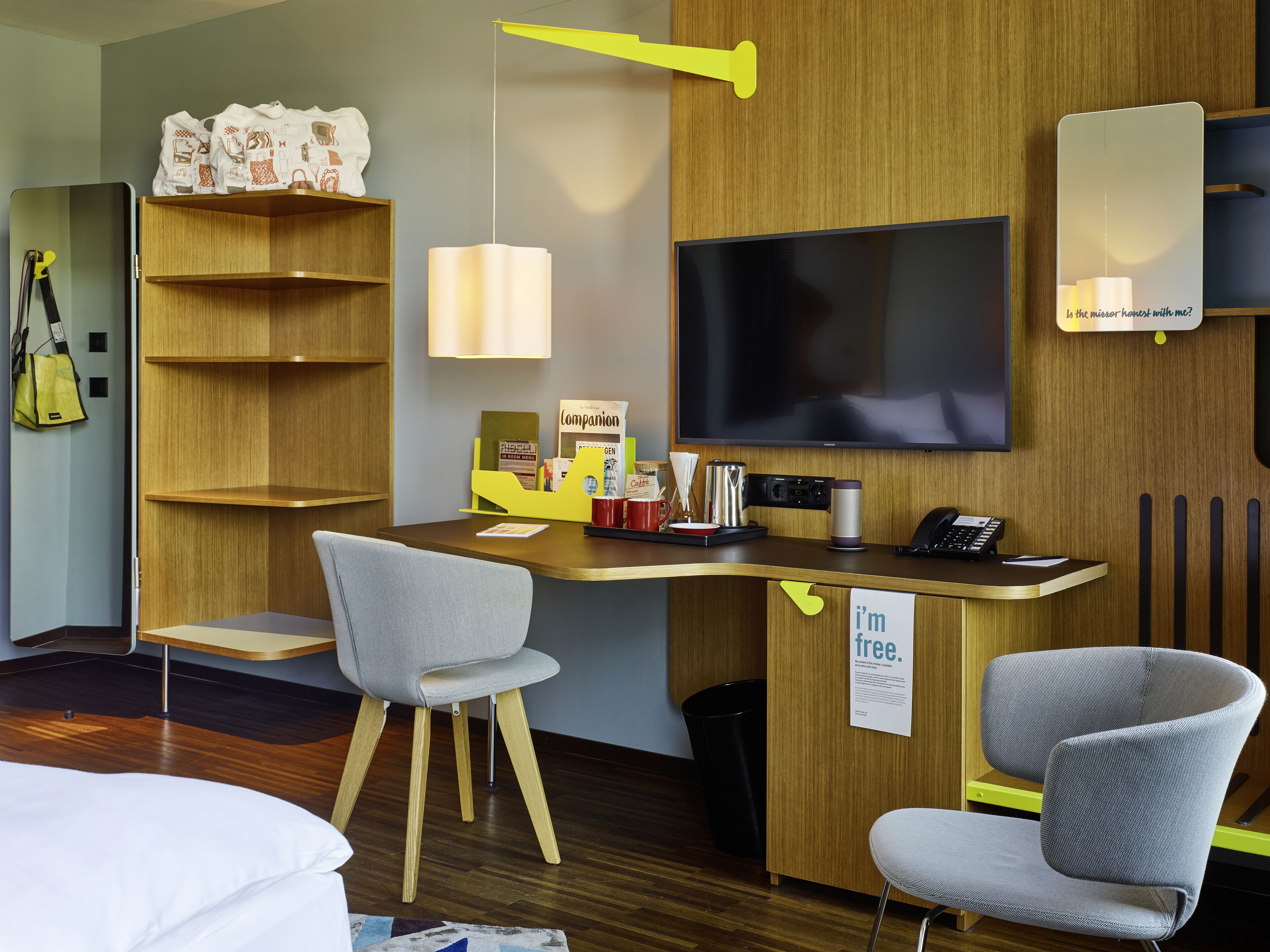 Una habitación de hotel moderna con un escritorio de madera, dos sillas grises, un televisor de pantalla plana, una estantería y un letrero que dice 'Estoy libre'.