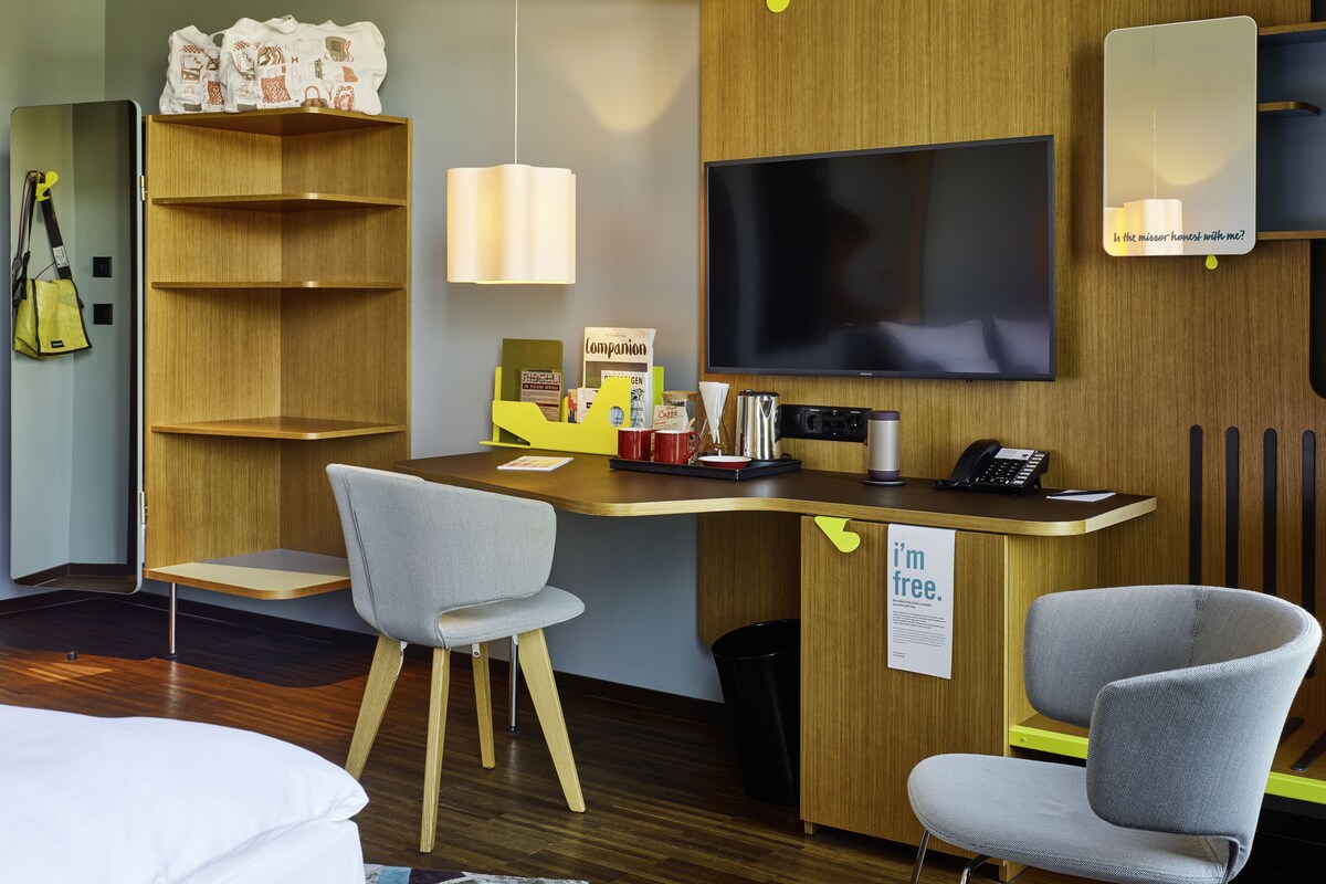Une chambre d’hôtel moderne avec un bureau en bois, deux chaises grises, un téléviseur à écran plat, une bibliothèque et un panneau indiquant : « Je suis disponible. »