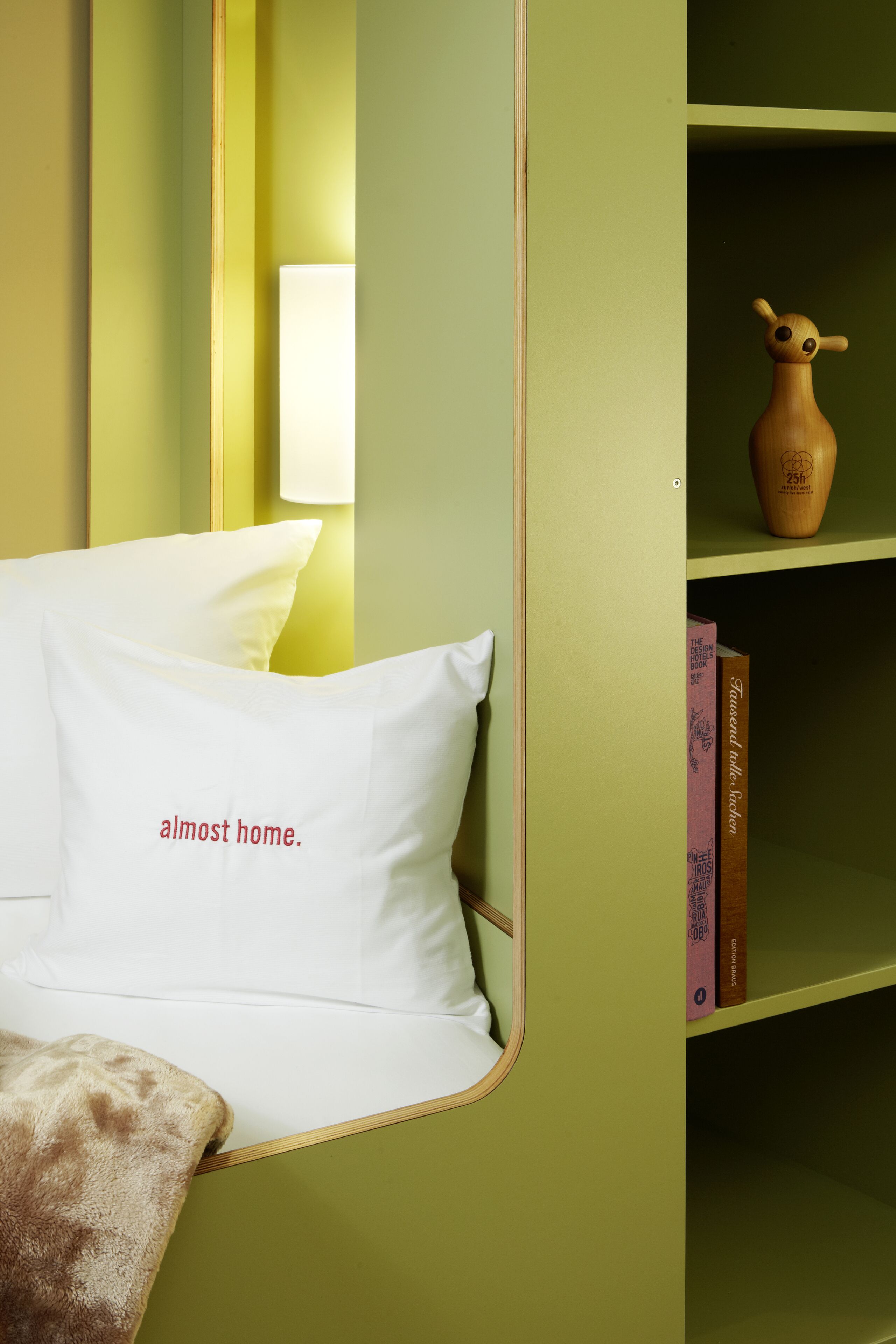 A room with a green wall, a white pillow with text, a lamp, and a bookshelf with books and a vase.
