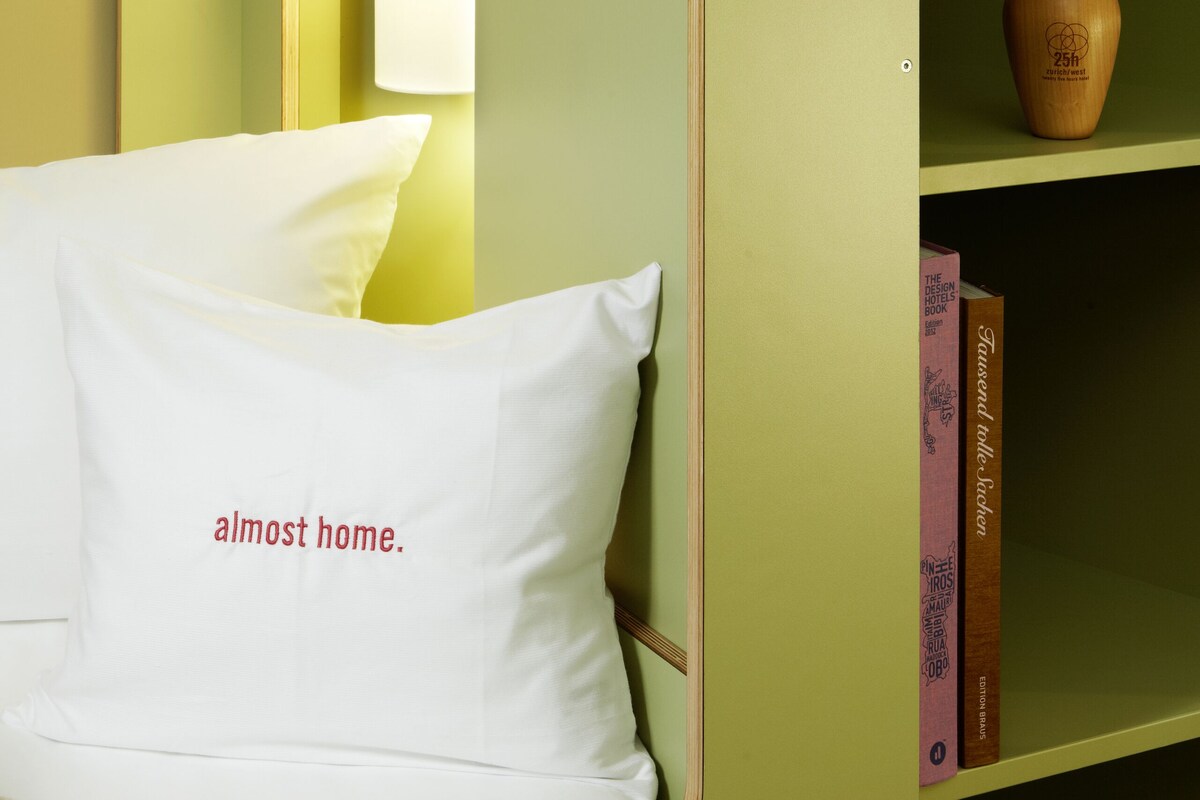 Une pièce avec un mur vert, un coussin blanc orné d’un motif, une lampe et une bibliothèque contenant des livres et un vase.