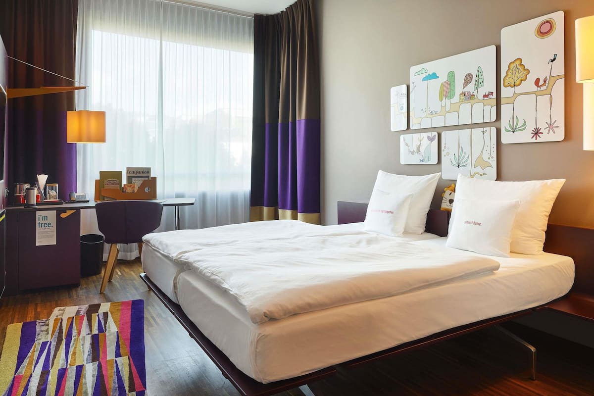 A hotel room with a large bed, a desk with a computer, a chair, and a desk lamp. There are curtains, a window, and wall art.