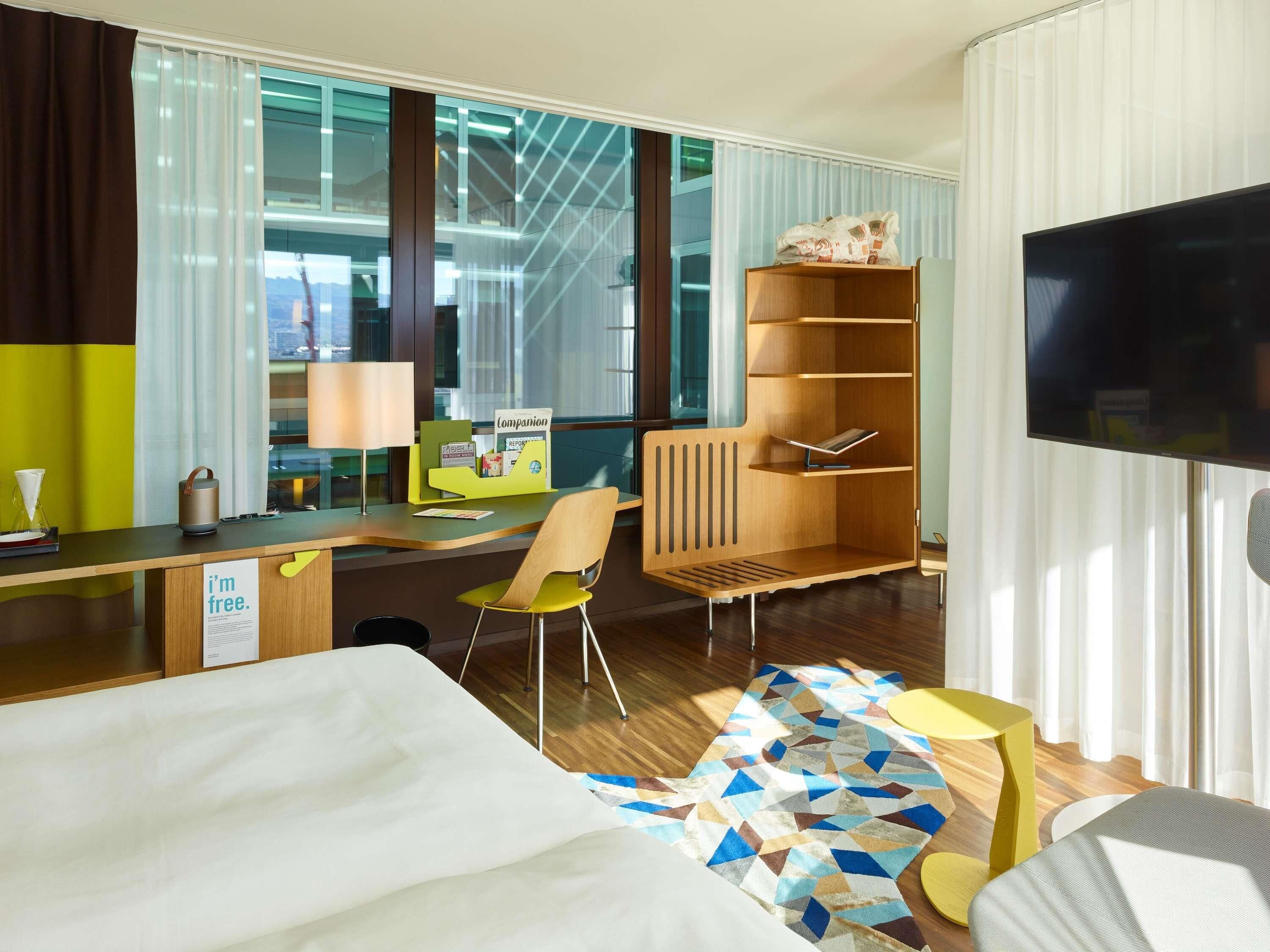 Una habitación de hotel con una cama, un escritorio, una silla, una estantería, un televisor y una ventana con cortinas.