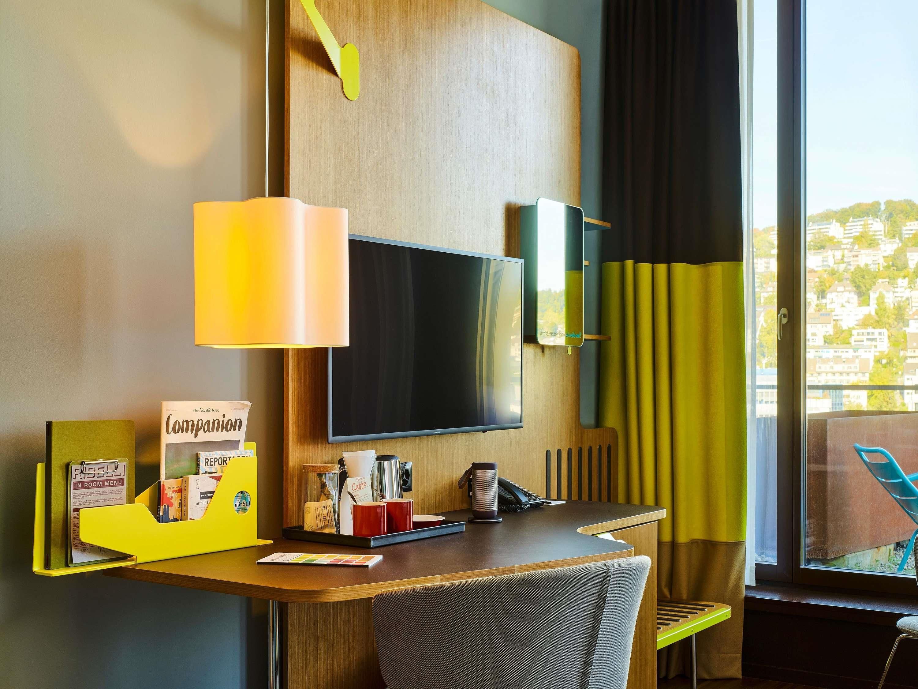 Ein Hotelzimmer mit Schreibtisch, Flachbildfernseher, Lampe, Stuhl, Fenster mit Vorhängen und Blick auf Gebäude.