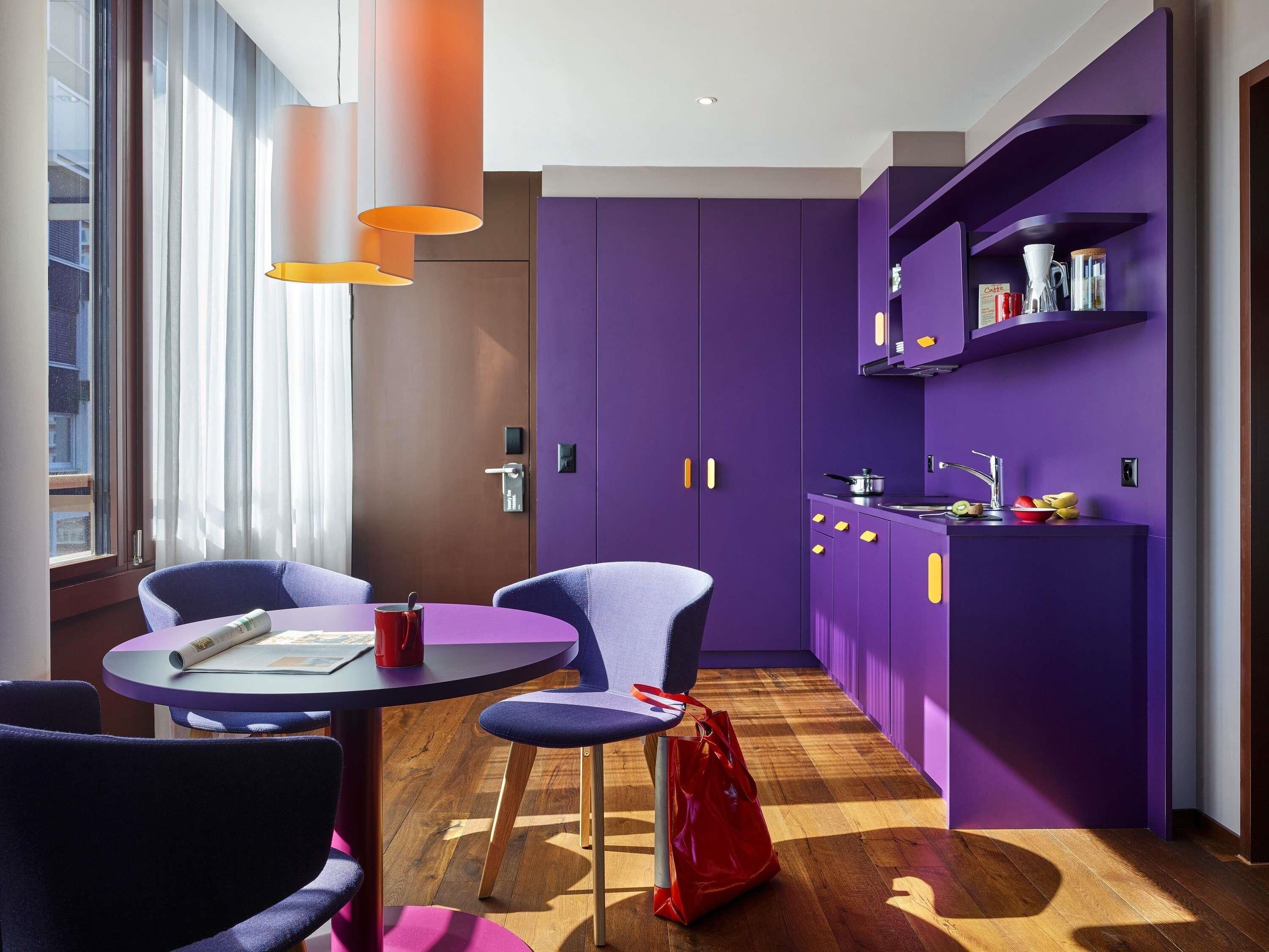 Een moderne keuken met paarse kastjes, een ronde eettafel met stoelen en een rode tas op de vloer.