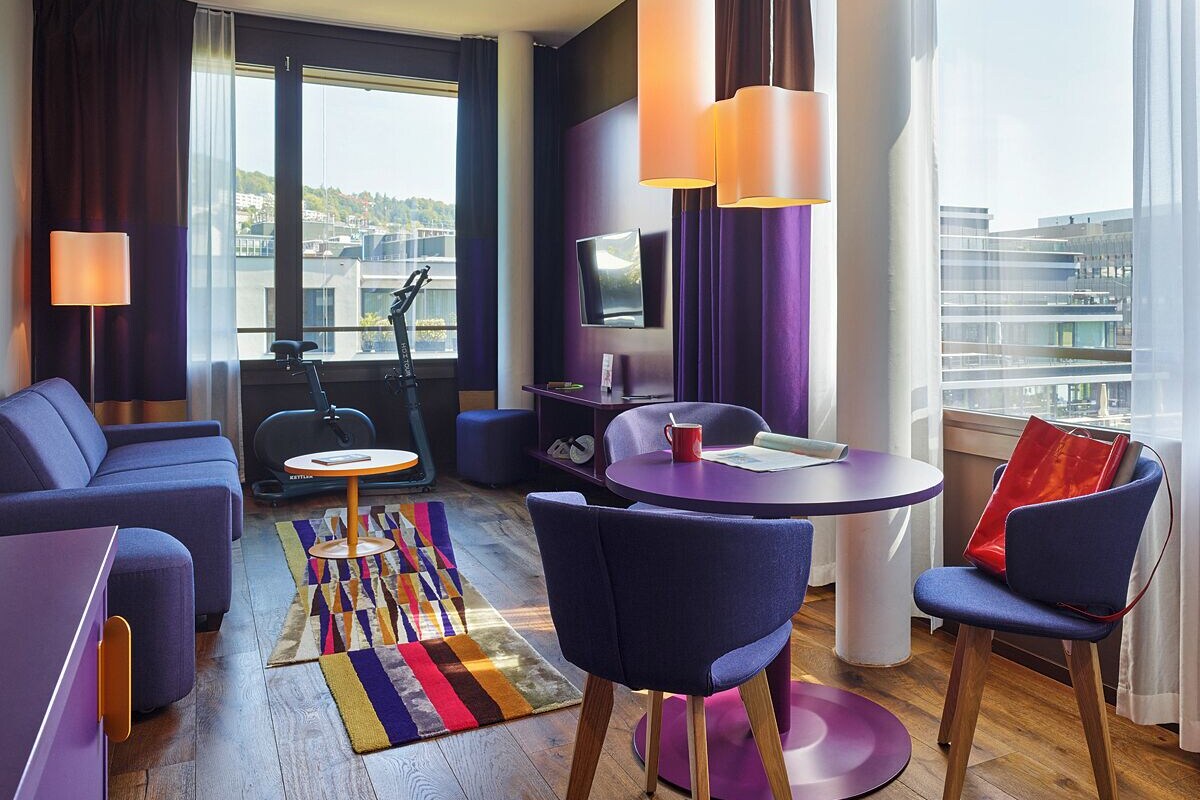 Une chambre d’hôtel moderne équipée d’un canapé, de fauteuils, d’une petite table, d’un tapis de course et offrant une vue sur la ville.